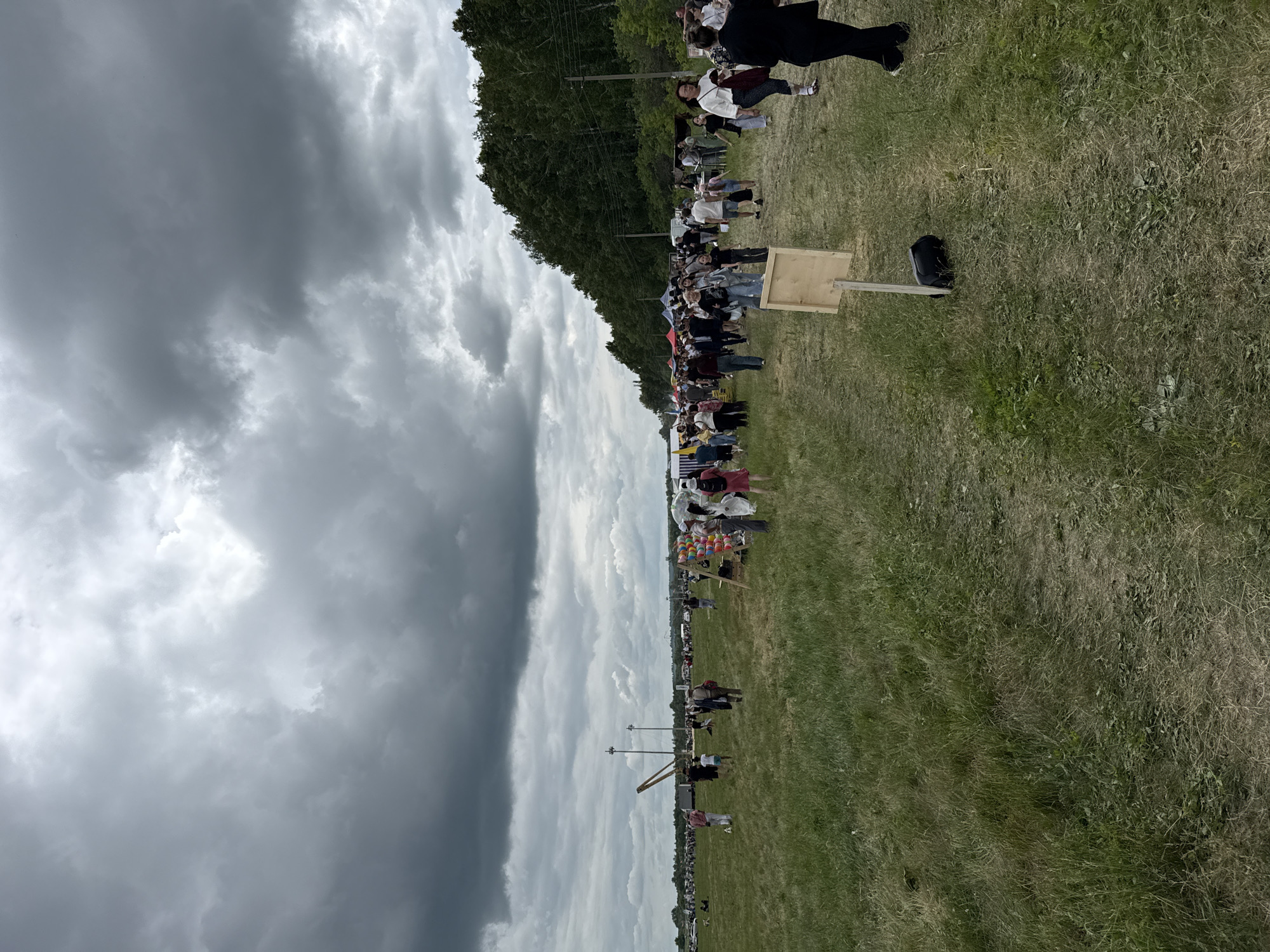 The image depicts an outdoor event with a crowd of people gathered in a grassy field under a cloudy sky. In the background, a dense line of trees forms a dark green border along the horizon. Several tents and stalls are visible among the crowd, suggesting a market or festival. People are seen walking, standing, and interacting. The ground is covered in green grass, with some areas appearing dry and sparse. There are also a few scattered objects, including a wooden signpost with a blank rectangular panel supported by a single wooden post and a black bag at its base. In the mid-ground, a tall, slender structure resembling a crane or gantry is present. The overall atmosphere suggests a daytime gathering, possibly in the city of Nurkeevo, Russia, given the context provided.