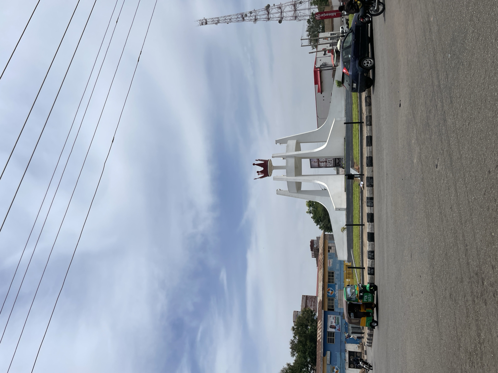 This rotated image captures a daytime street scene in Jos, Nigeria, featuring an urban landscape with a central monument, vehicles, and buildings under a partly cloudy sky.
In the middle ground, a large, white, angular monument, possibly a roundabout fixture, stands prominently. It is topped with a distinctive red, crown-like sculpture. A sign on the monument clearly displays the text "HE IS RISEN". The monument is situated beside a paved road, which features a curb with black and white stripes.
On the road, a dark blue hatchback car is visible, and further down, a green Keke Napep (auto-rickshaw) with "PTA 11250" written on its side is parked. A motorcycle can also be seen near the auto-rickshaw.
Along the left side of the road, a row of buildings with blue-painted facades is visible. Signage on these buildings includes "StarTimes" and a political poster with "2023" and "SIMON" partially legible. In the background, a tall, metallic communication tower with numerous dishes and antennas rises high into the sky. Another distant building features a banner with the text "Kilimanjaro" partially visible.
The sky is bright with scattered white clouds, suggesting a clear day with good visibility. Multiple overhead power lines stretch diagonally across the upper left quadrant of the frame. The scene portrays a typical urban environment with daily street activity.