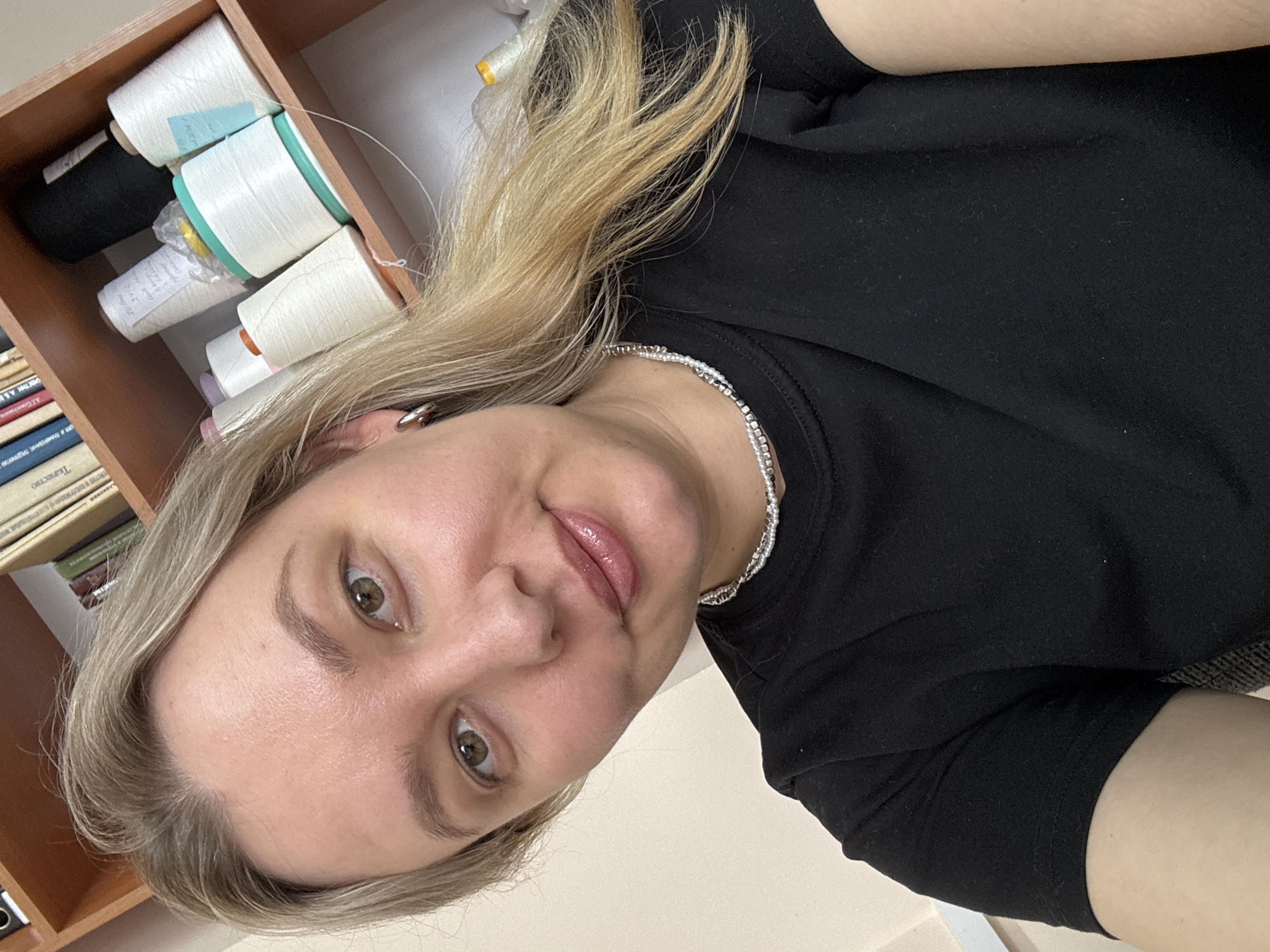 A selfie depicts a woman in a black t-shirt with light-blonde hair and a silver necklace, indoors. Behind her is a wooden shelf containing spools of thread in varying colors (white, black, etc.), and books. The exact location in the city of Chaikovsky, Russia, is not discernible from the image.