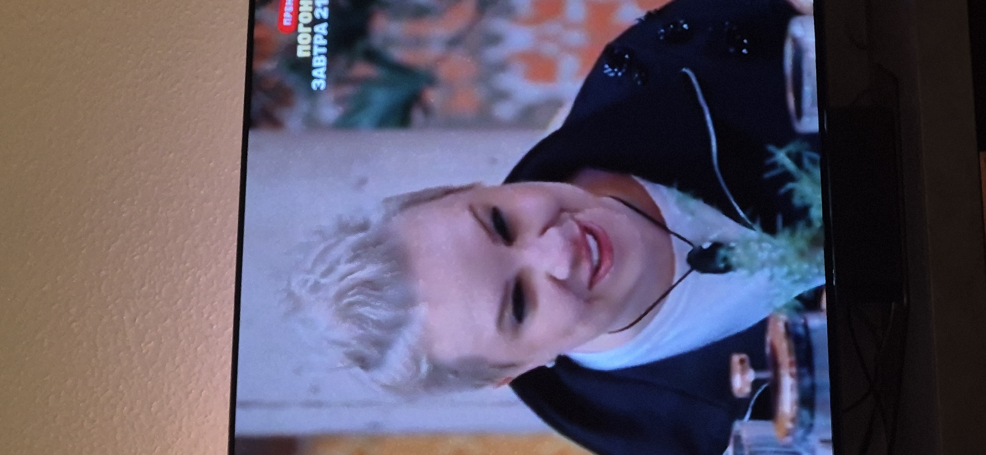 The image shows a medium shot of a woman with short, spiky blonde hair. She is wearing a white t-shirt and a black jacket with beading on the lapels. She has dark eyeliner and is smiling, revealing her teeth. A dark pendant necklace is visible on her chest, and a sprig of greenery is also present near her neck. In the background, there is a blurred brick pattern and some text that appears to be a television broadcast overlay, with the words "ПРЕМЬЕРА" (Premiere) and "ЗАВТРА 21" (Tomorrow 21) visible. The scene is indoors, and the lighting suggests it is a staged setting, possibly a television studio or a recorded broadcast. There is no information in the image to suggest a location in Baden-Baden, Germany.