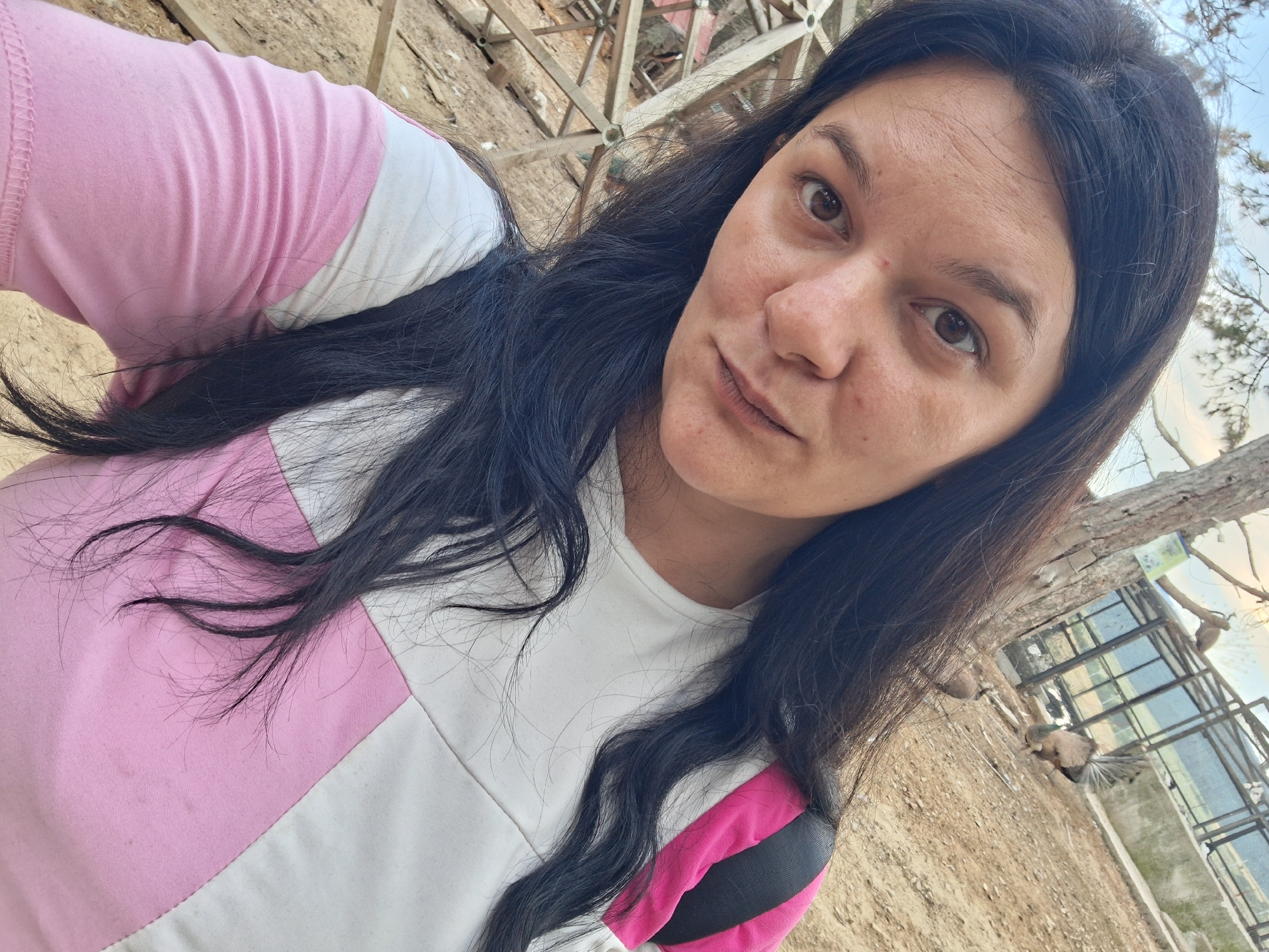 In the city of Hadzhidimovo, Bulgaria, a woman with long, dark hair is captured in a close-up selfie. She is wearing a white t-shirt with large pink and white geometric panels and a black strap visible across her shoulder. Her gaze is directed towards the camera, and her expression is neutral. The background suggests an outdoor environment, possibly a park or zoo, with hints of wooden structures and fencing. In the distance, behind a fence, two birds, one with its tail feathers fanned out, are visible. The ground appears to be dirt or gravel. The lighting indicates it is daytime.