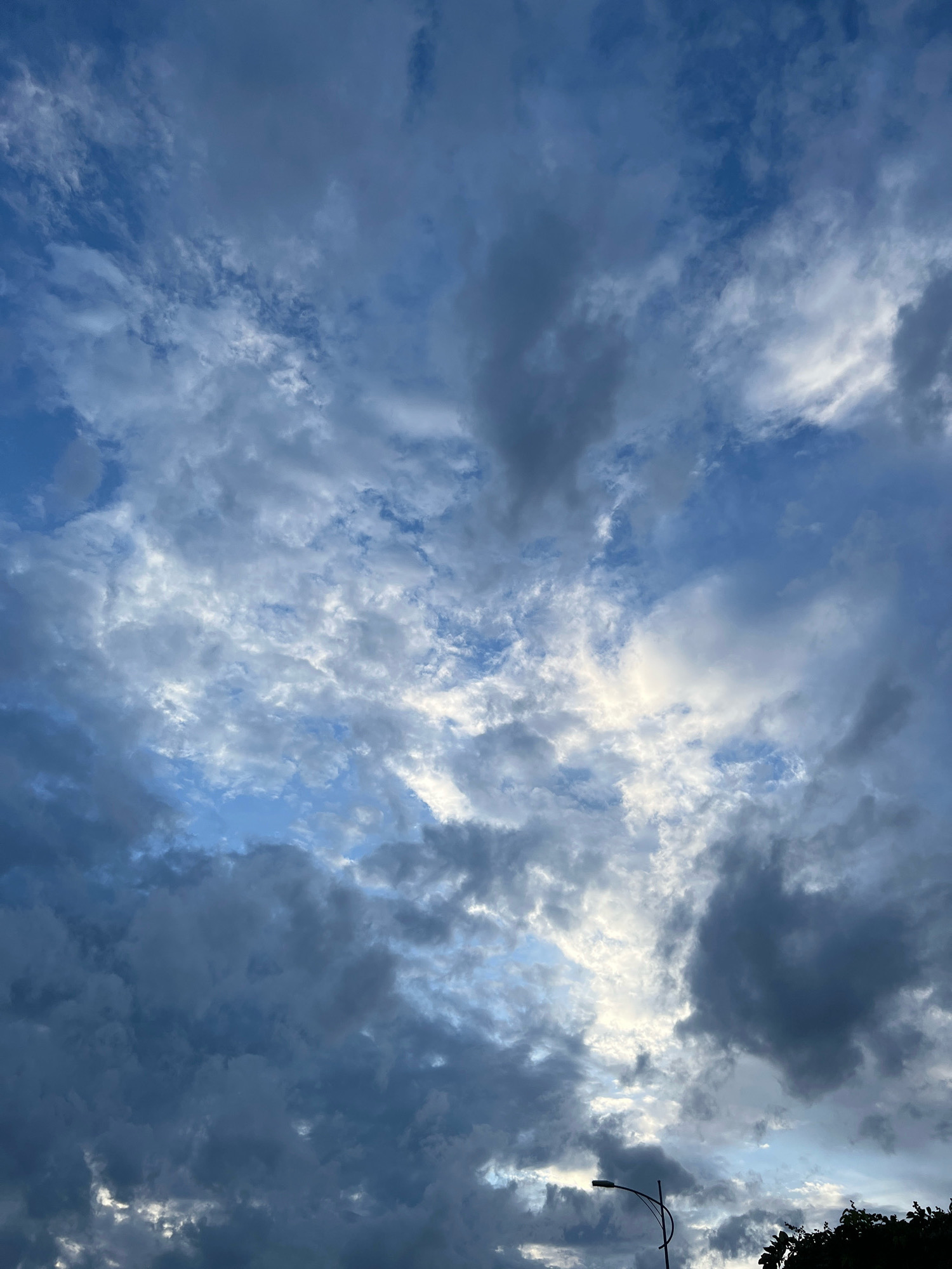 This is a vertical, eye-level, outdoor shot of a cloudy sky over Nha Trang, Vietnam. The clouds are varied, with some dark and heavy, and others lighter and more scattered. A few patches of blue sky are visible between the clouds. A street light is visible in the lower right portion of the image, and the top of a tree is also visible at the bottom right. The lighting suggests it may be dusk or dawn, or possibly an overcast day. There are no people or specific landmarks visible that would definitively place it within Nha Trang city, beyond the general atmospheric conditions of a coastal Vietnamese location.