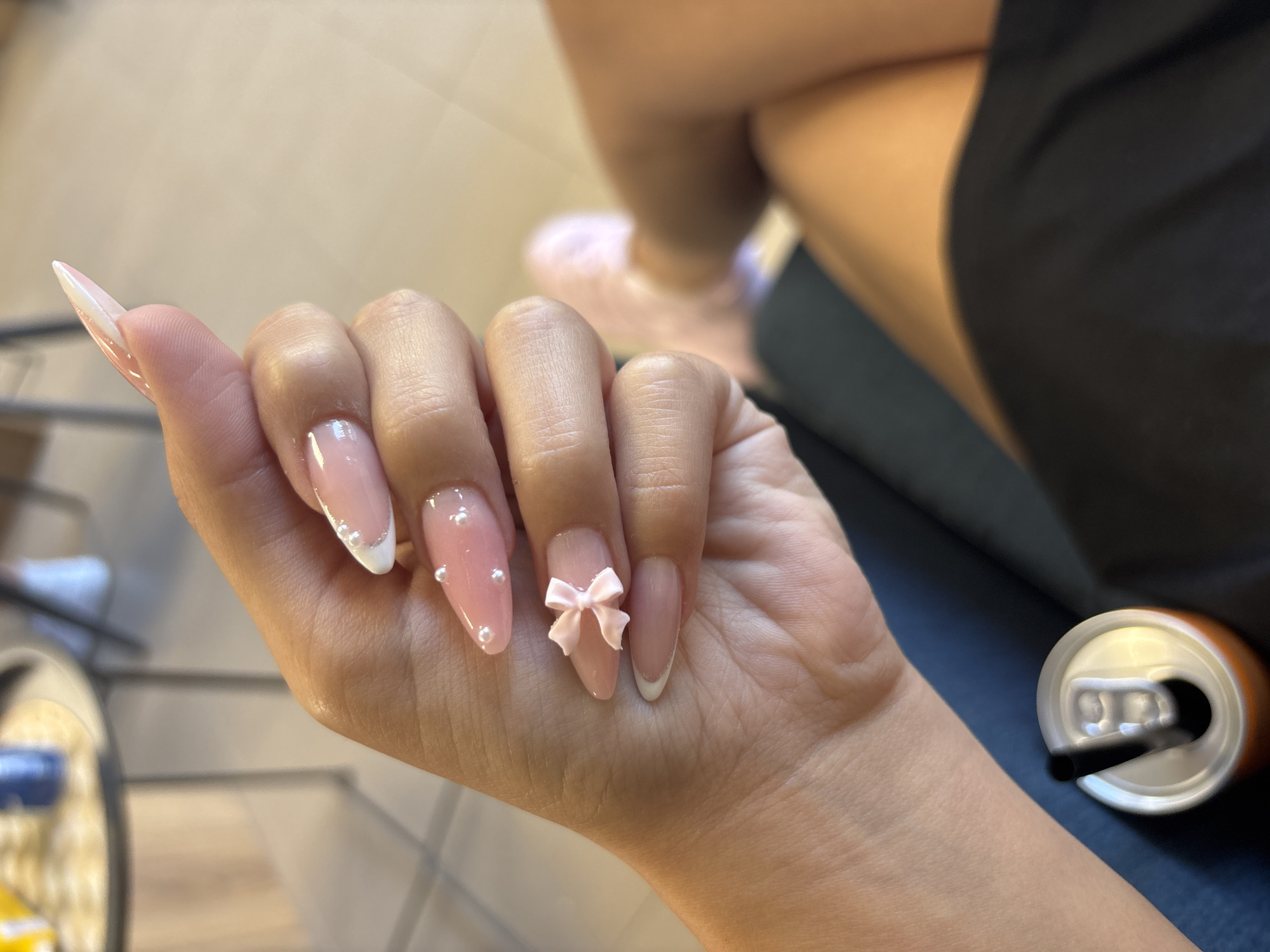 A woman's hand with a pink French manicure and pearl embellishments is held up in front of her. Her nails are long and almond-shaped. One nail has a small pink bow charm. She is wearing a dark blue top and black shorts. In the lower left of the image, a can of soda with a black straw is visible. The background is blurred but appears to be an indoor setting, possibly a cafe or salon. There are no discernible landmarks that would indicate a specific location in Plovdiv, Bulgaria.