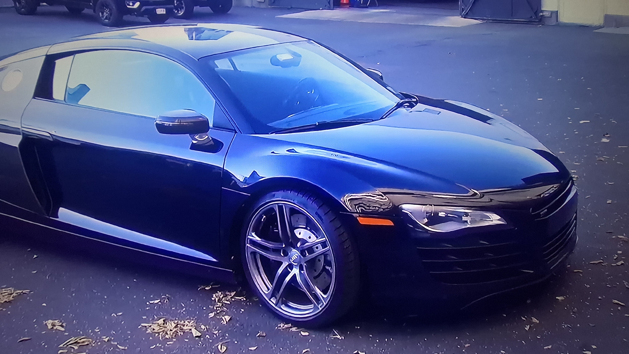 A dark blue Audi R8 is parked on an asphalt surface, littered with small piles of brown leaves. The car is positioned at a slight angle, with its front end facing towards the right of the frame. The car's silver rims are visible, along with a portion of its front grille, headlights, and side mirror. In the background, partially obscured, are two dark colored pickup trucks. A light colored building is visible behind the vehicles in the background. The location is a parking lot in an urban area.