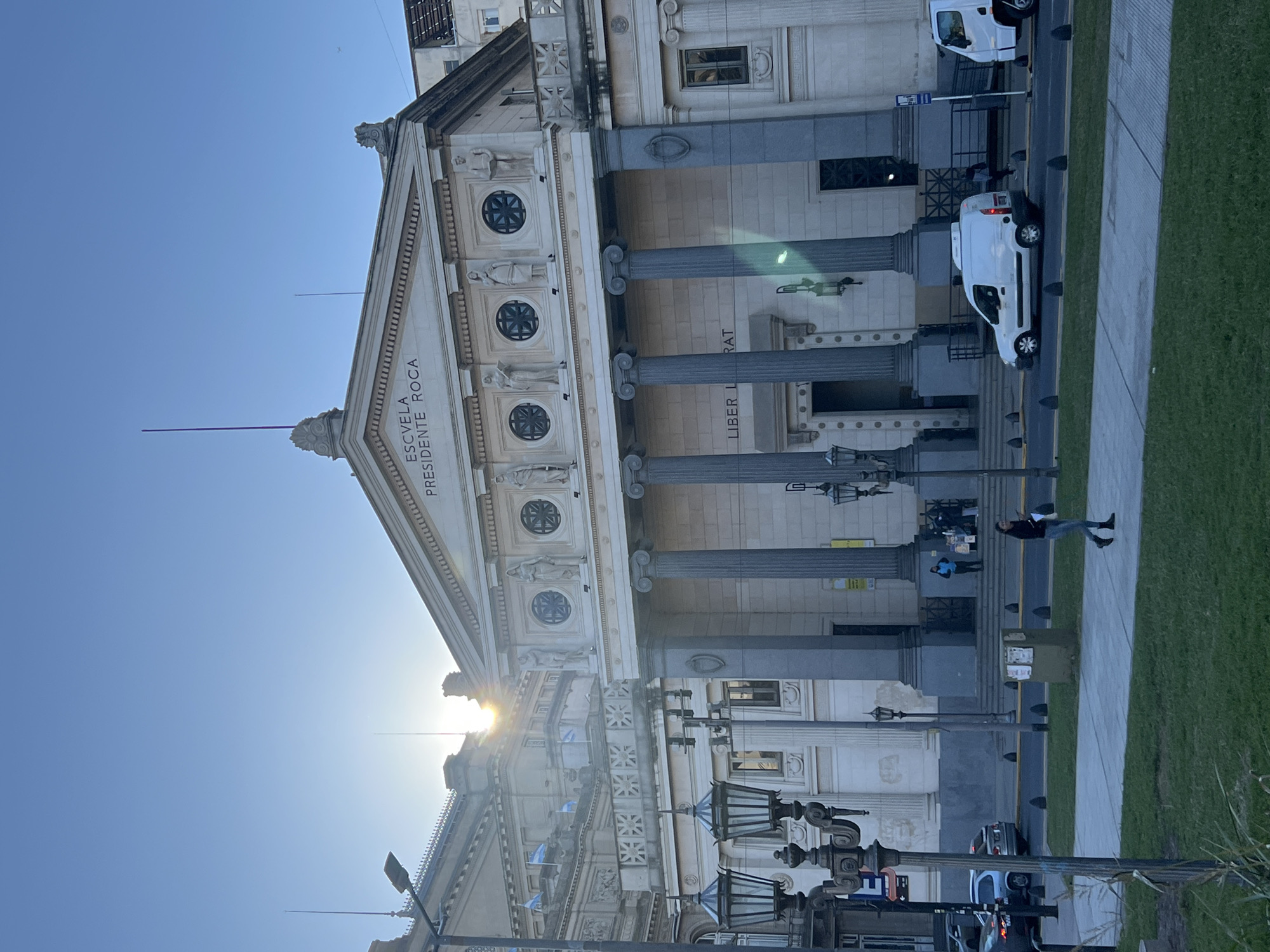 The image captures the Escuela Presidente Roca, a historical school building in Buenos Aires, Argentina. The building features a neoclassical facade with columns and ornate detailing, including the inscription "ESCUELA PRESIDENTE ROCA." The image is taken at street level, looking upward at the building. In the foreground, there are antique-style street lamps. Cars and white vans are parked on the street alongside a sidewalk with pedestrians. One person is seen lying on the sidewalk. The sun is positioned behind the building, creating a bright glare. The sky is clear and blue.