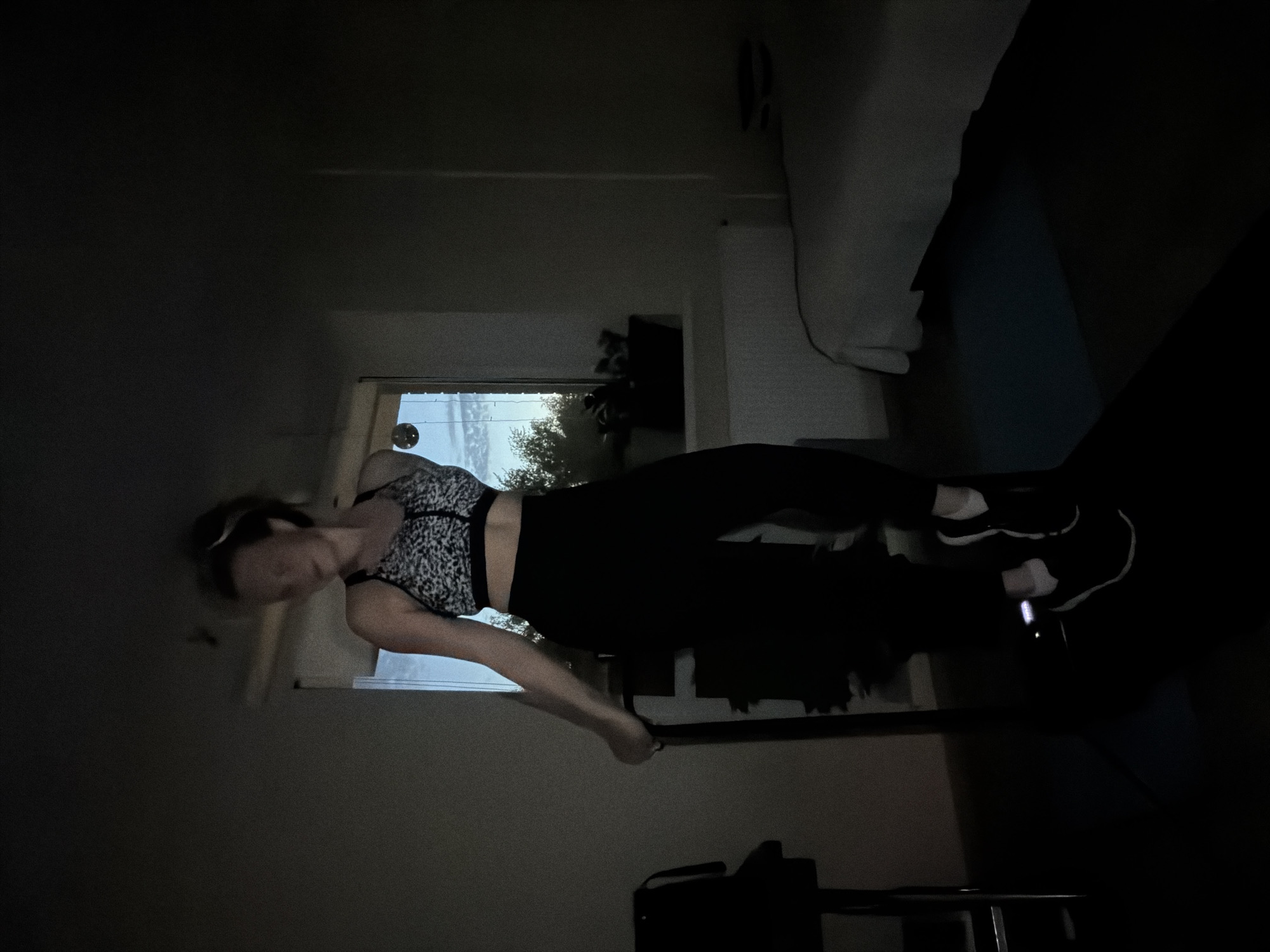 The image, rotated 90 degrees clockwise, captures a young woman engaged in a workout session indoors, likely in a bedroom or home gym in Izhevsk, Russia. The room is dimly lit, with natural light from a large window behind her providing the primary illumination.
The woman is dressed in a black and white leopard-print sports bra, black athletic leggings, and black sneakers with white accents. Her hair is pulled back, and her face is slightly blurred, suggesting movement. She stands with her body slightly angled, appearing to be in the midst of an exercise. Her right arm extends downwards, seemingly holding a black strap or handle, possibly part of a resistance band or attached to a small piece of workout equipment like a dumbbell visible on the floor near her feet. Her left arm is bent, holding a small dark object, possibly a phone. She appears focused and concentrated on her activity.
Through the window behind her, a bright daytime scene is visible, featuring green trees against a light, possibly overcast, sky. The interior details include a bed with white bedding visible in the upper right background. The overall atmosphere suggests a quiet, personal workout environment. There is no visible text.