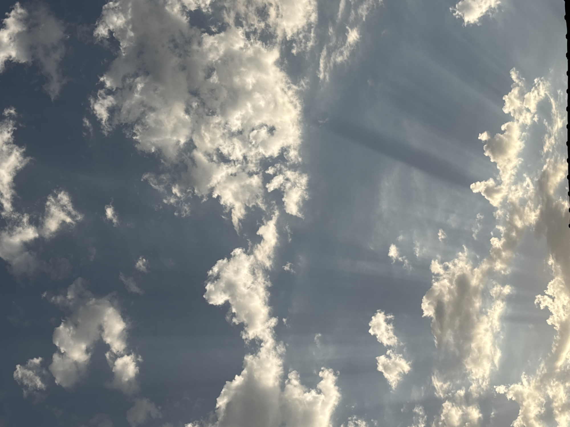 The image is a vertical, full-frame shot of a cloudy sky, likely taken from ground level. The sky is a mix of blue and grey, with scattered white clouds. Sunbeams are visible, creating a dramatic lighting effect. The bottom edge of the frame shows a dark, silhouetted object, possibly a rooftop or a line of trees, with some faint illumination on its upper edge. The scene is devoid of any identifiable landmarks or people, making it impossible to pinpoint a specific location within Taraz, Kazakhstan. The overall impression is one of a natural atmospheric phenomenon.
