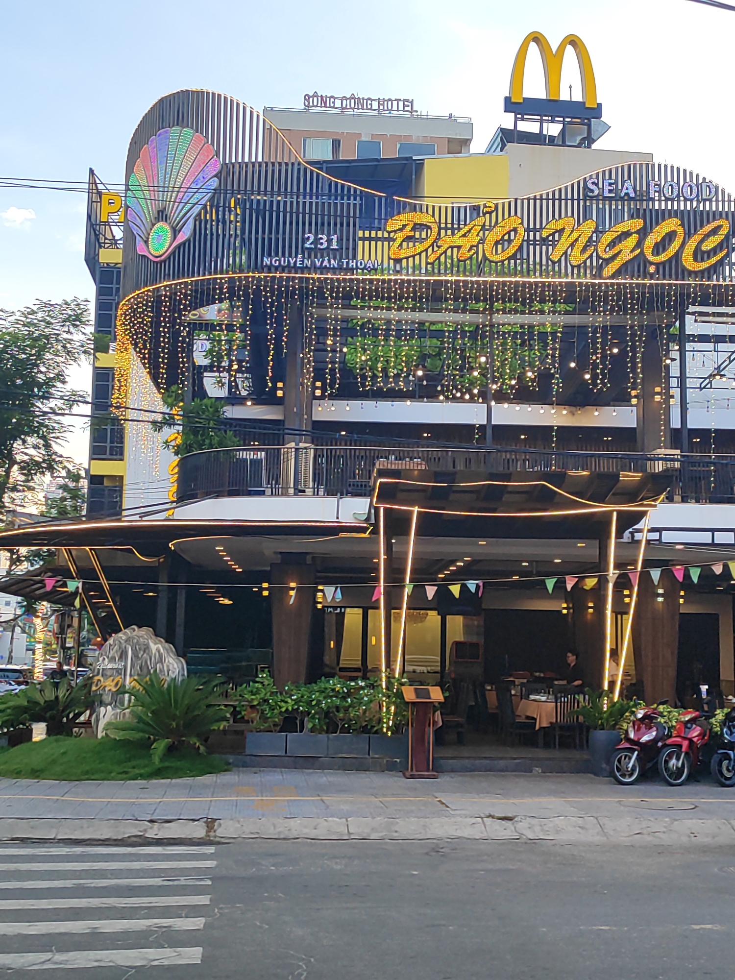 The image captures a street-level view of a brightly lit restaurant exterior in Đà Nẵng, Vietnam. The restaurant, named "ĐẢO NGỌC" and identified as "SEA FOOD," is adorned with numerous string lights and colorful banners. A prominent large shell-shaped logo with a pearl is displayed on the building's facade, alongside the street address "231 NGUYỄN VĂN THOẠI". Above the restaurant, the golden arches of a McDonald's sign are visible, indicating proximity to the fast-food chain. In front of the restaurant, a stone-like structure with the restaurant's name inscribed is surrounded by lush greenery. The sidewalk is paved with gray tiles, and a pedestrian crossing is marked on the asphalt street. Several motorcycles are parked along the sidewalk. Two people are visible near the entrance of the restaurant. The sky is clear and bright.