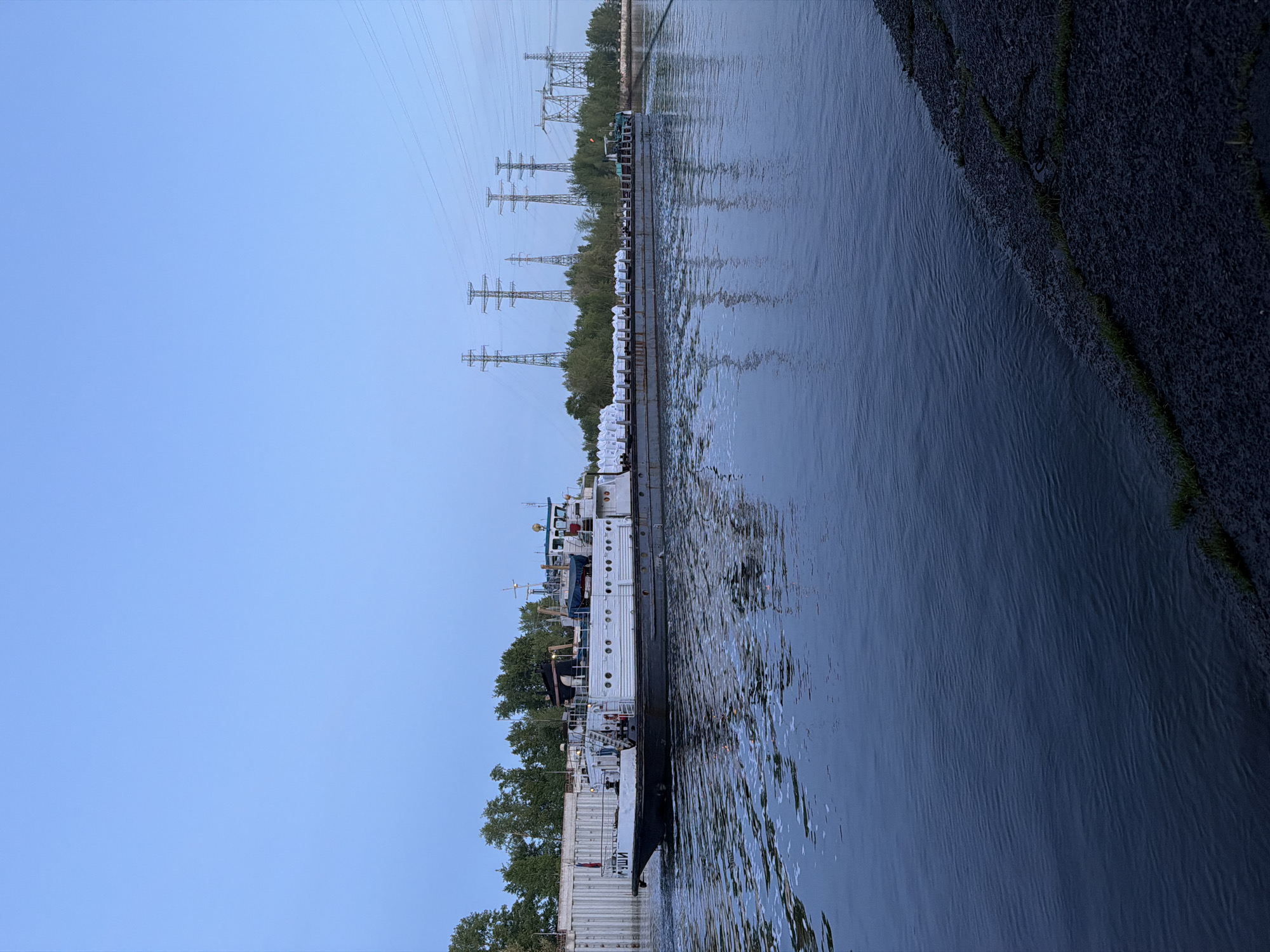 A river view in the city of Chaikovsky, Russia, featuring a barge loaded with white, possibly sand-filled, bags docked along a concrete riverbank lined with trees. Alongside the barge is a multi-tiered vessel. Overhead, power lines extend across the river's edge. The opposite bank is visible in the distance, obscured by trees and other vegetation. The sky is a clear, pale blue. A dark, rocky embankment borders the river on the right side of the image. The water shows slight ripples, indicating a calm surface.