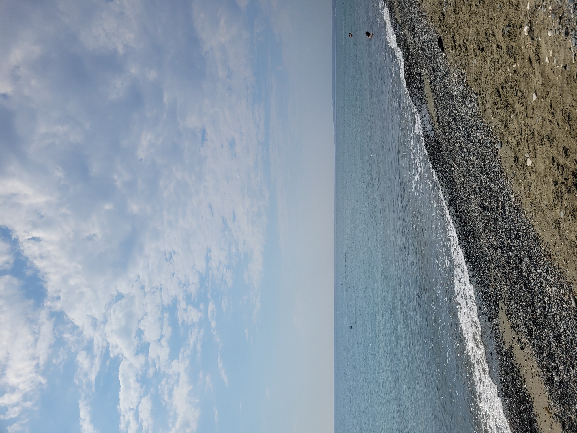 The image depicts a beach scene under a partly cloudy sky. The foreground features a sandy beach transitioning into a pebble-covered shoreline. Gentle waves with white foam are breaking on the shore. The sea is a calm, blue expanse stretching to the horizon. In the distance, the hazy outline of land is visible. A few people are in the water, with one individual appearing to swim. A white buoy is also visible in the sea. The overall atmosphere is serene and suggests a day at the beach.