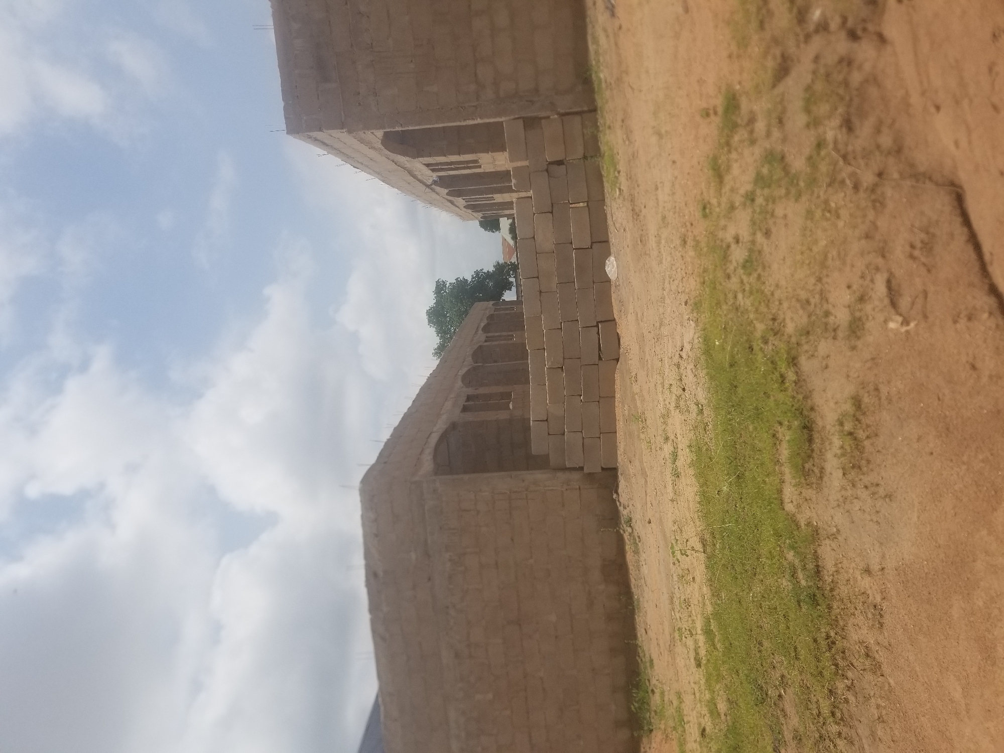 In the city of Jauro Yinu, Nigeria, a construction site is captured under a cloudy sky. The scene focuses on two unfinished brick buildings. The building on the left is a solid, long structure with arched window openings and a slightly sloped roofline. In front of it, a raised foundation or stairway made of stacked concrete blocks is visible. To the right, another building is under construction, also with arched openings and a higher roofline, indicating it may be taller or at a later stage of development. A pile of concrete blocks lies in the foreground between the two structures. The ground is a mix of dirt and sparse green grass, suggesting an outdoor, undeveloped area. The overall impression is one of ongoing construction in a rural or semi-urban Nigerian setting.