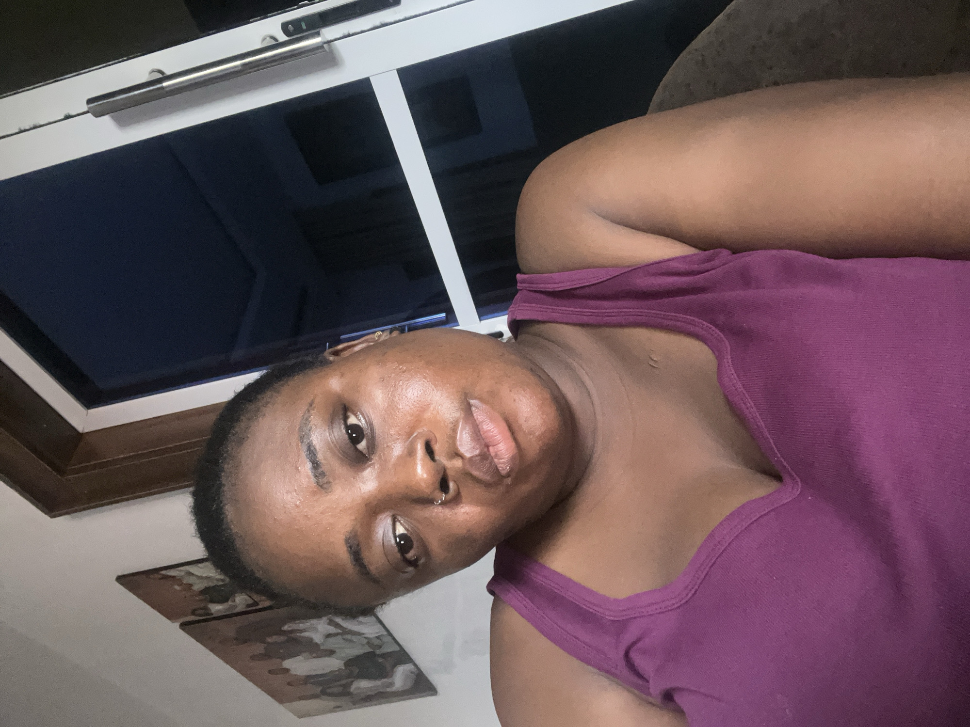 A person is wearing a purple tank top and is lying down. Behind the person is a window with a silver handle. On the wall, there is a framed picture. The location is not specified in the image. Therefore, the location in the city of Jos, Nigeria is unknown.