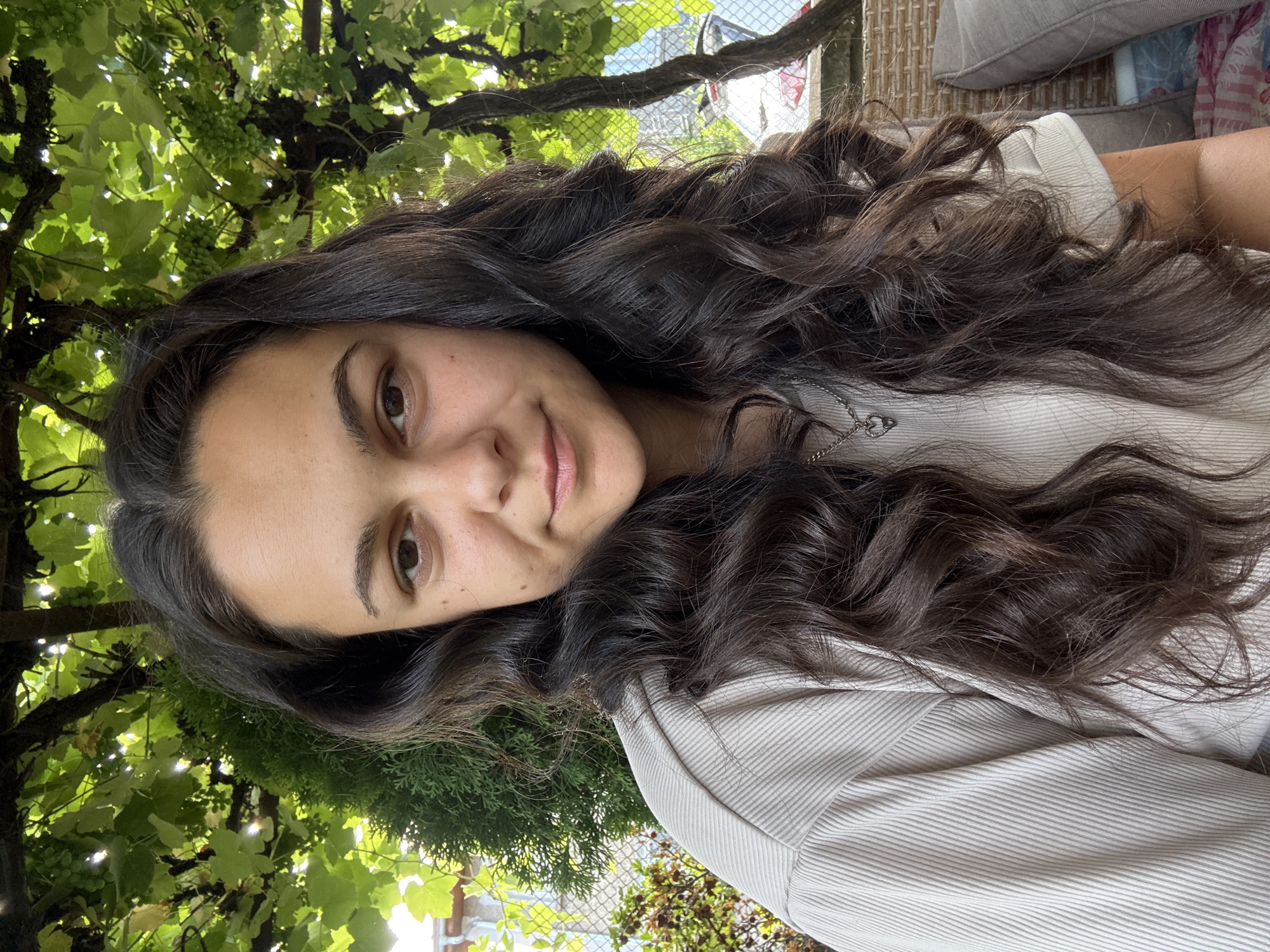 A young woman with long, dark, wavy hair is pictured from the chest up, outdoors. She is wearing a light-colored, ribbed shirt and a silver necklace with a heart pendant. The background consists of green foliage from a grapevine and a coniferous tree. Behind the foliage, a light-colored fence and a glimpse of a building are visible. The woman is looking towards the camera with a slight smile. The location is identified as Oreshak, Bulgaria.