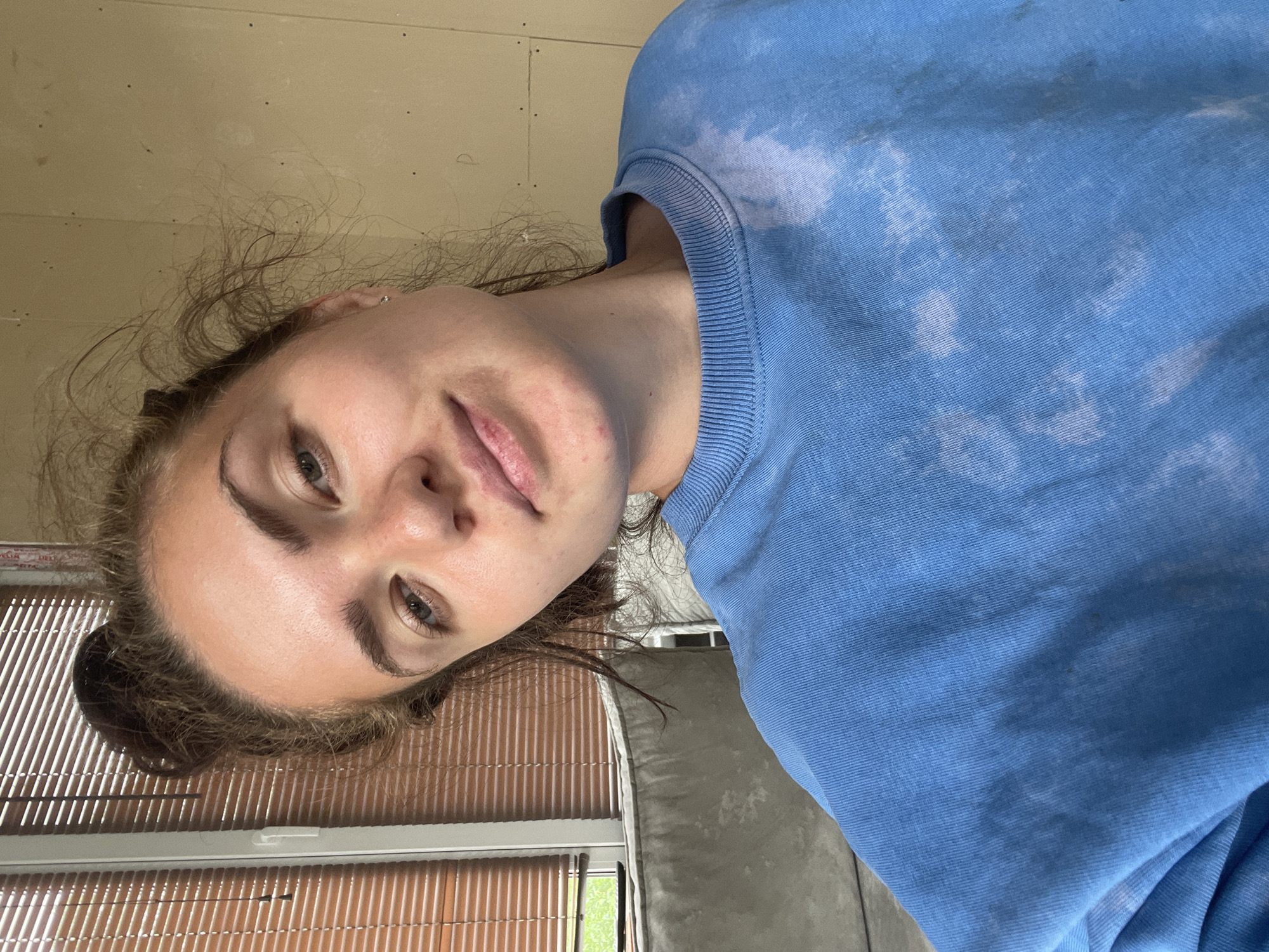 A young woman with her hair pulled up in a messy bun is looking at the camera. She is wearing a blue sweatshirt with a bleached pattern. Her face is angled to the left. In the background, there is a window with blinds and a grey sofa. The wall behind her is a light beige color. There is no indication of the specific location within Мосметаллоконструкция Онт, Russia.