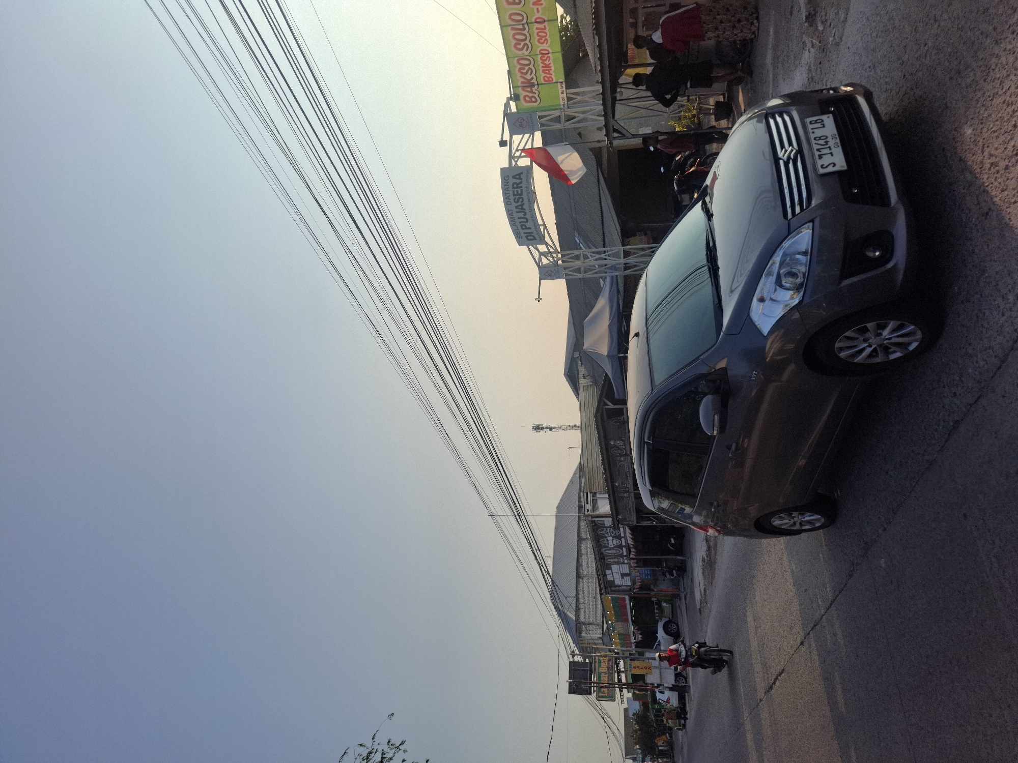 The image depicts a street scene in Sumberwudi, Indonesia, during what appears to be late afternoon or early evening due to the low angle of the sun. The sky is a clear, pale blue, with numerous electrical wires crisscrossing overhead, indicating an urban or semi-urban setting.
In the foreground, a grey Suzuki Ertiga car is parked on the right side of the road, facing away from the camera. Its license plate reads "S 1148 LB". The car is casting a long shadow onto the asphalt road, which is marked with a white line down the center.
To the left of the car, a motorcycle is being ridden by a person wearing a red shirt and a helmet, moving away from the camera down the road. On the left side of the road, several shops are visible, with awnings and signage. One sign reads "Layar Buah". Further down the street, white cars are parked.
In the background, an archway with the text "SELAMAT DATANG DI PUJASERA" (Welcome to Pujasera) is prominently displayed, with an Indonesian flag hanging from it. To the right of the archway, a food stall is set up with a green banner that says "BAKSO SOLO" (Solo Meatballs). Several people are gathered around the food stall, some standing and some appearing to be customers. One person is wearing a red and white top, and another is wearing a patterned skirt. A tall antenna is visible in the far background, suggesting infrastructure in the vicinity. The overall environment appears to be a commercial area with food vendors and local businesses.