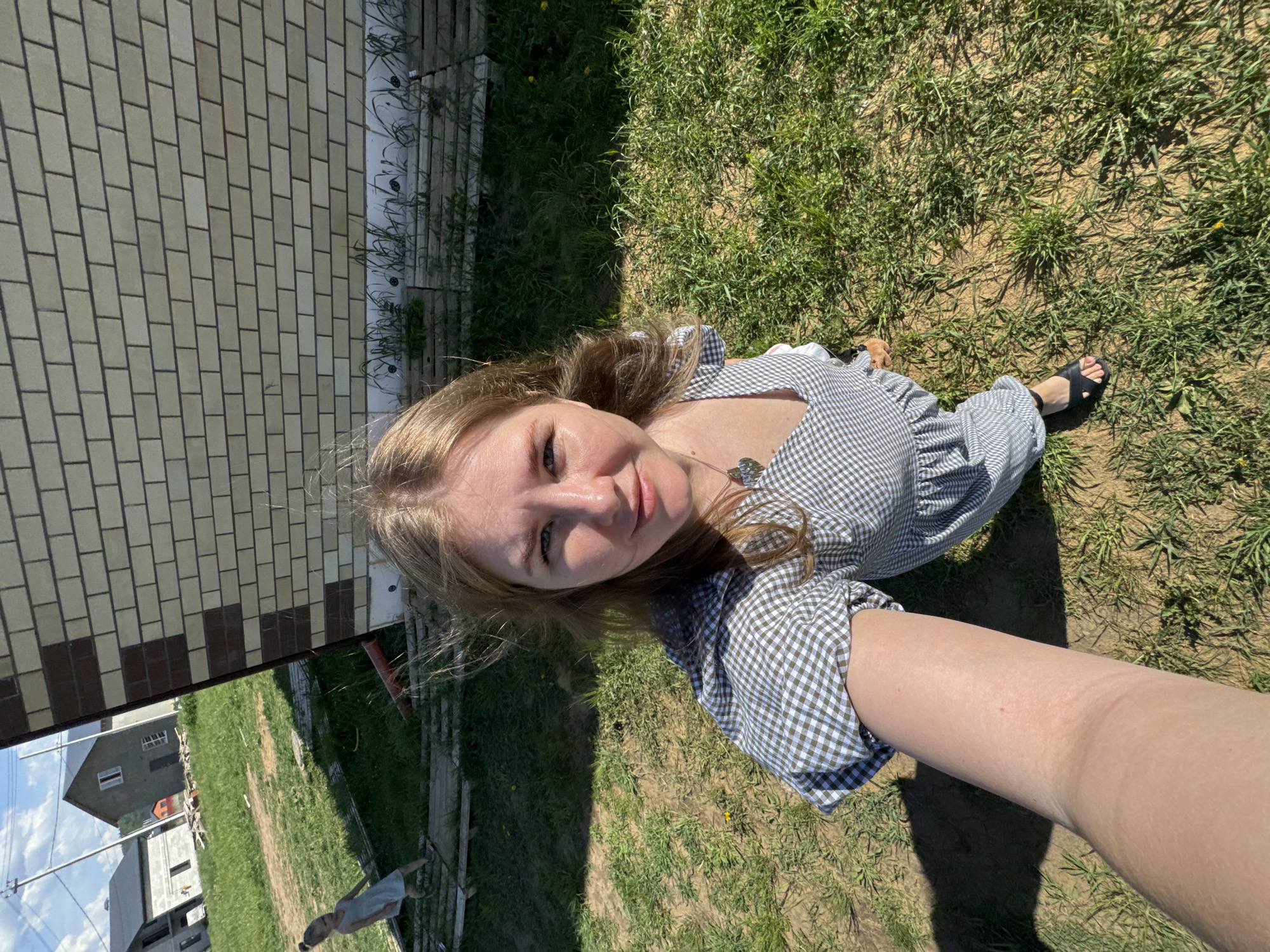 A woman lies on a patch of grass adjacent to a brick building in Ферма, Russia, looking up at the camera. She wears a checkered dress, a necklace, and sandals. The location includes grass, the brick structure, and in the background, several houses.