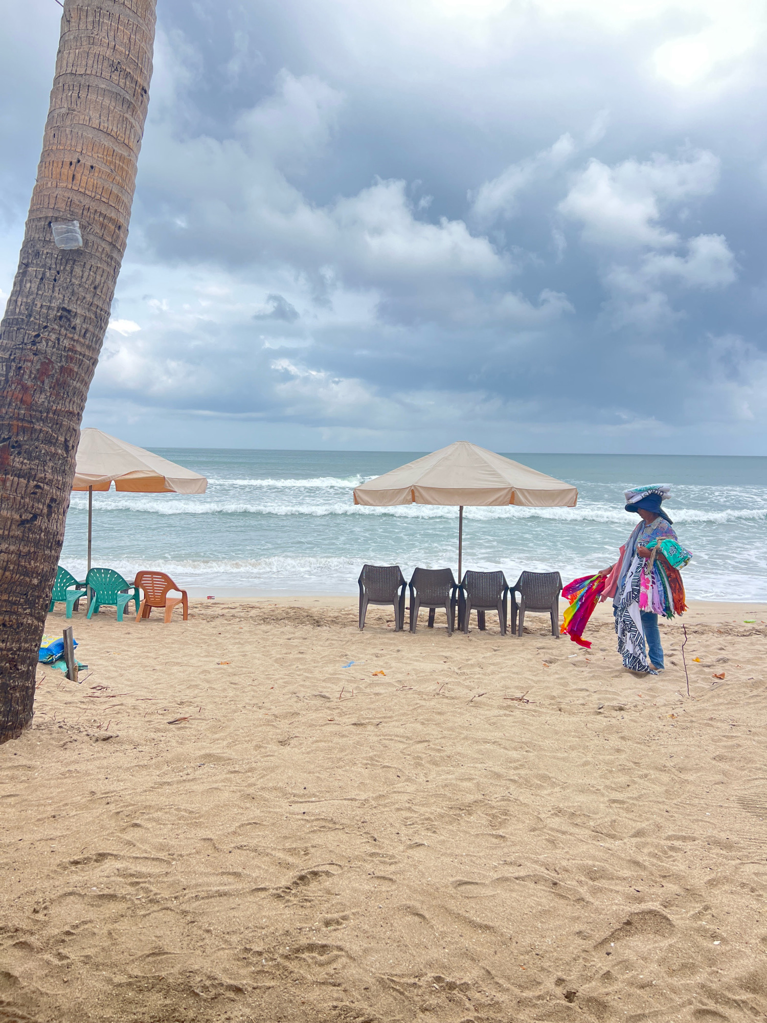 On a cloudy day at Kuta Beach, Bali, Indonesia, a person is standing on the sandy shore, holding several colorful fabrics. The person is wearing a blue hat, a patterned top, and jeans. To the left of the person are four empty plastic chairs and a large, light-colored beach umbrella. Further to the left, a palm tree trunk occupies the foreground. In the background, the ocean waves crash onto the shore under a dramatic, grey sky. Scattered pieces of trash are visible on the sand.