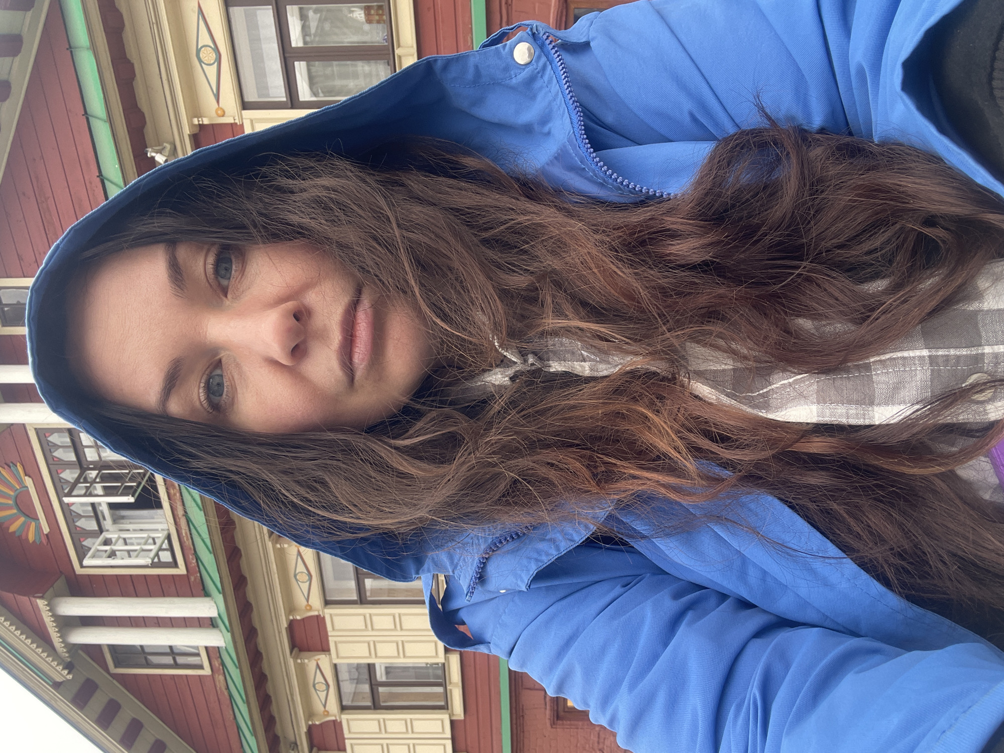 A woman is posing for a selfie. She is wearing a light blue hooded jacket. Her long brown hair is cascading around her face and over her jacket. Behind her is a colorful building with decorative trim, indicative of Russian architecture. Based on the architecture, the location is most likely in the city of Kazan, Russia.