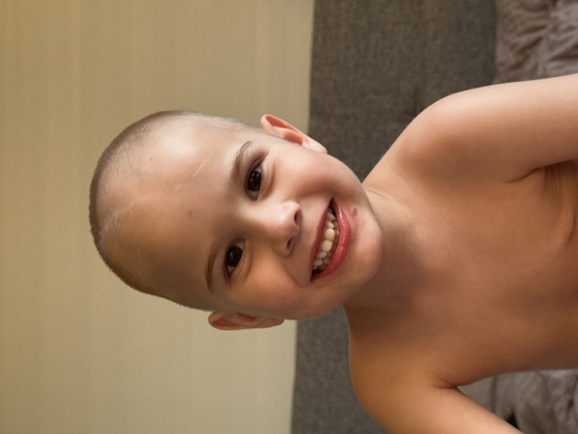 A young, fair-skinned child with a shaved head is smiling and looking directly at the viewer. The child is shirtless, and the background is composed of a pale wall on the left and a dark gray textured surface (possibly a couch or wall covering) on the right. The image appears to be taken indoors, and the lighting is soft. No other details are provided to conclude a location in the city of Chaikovsky, Russia.