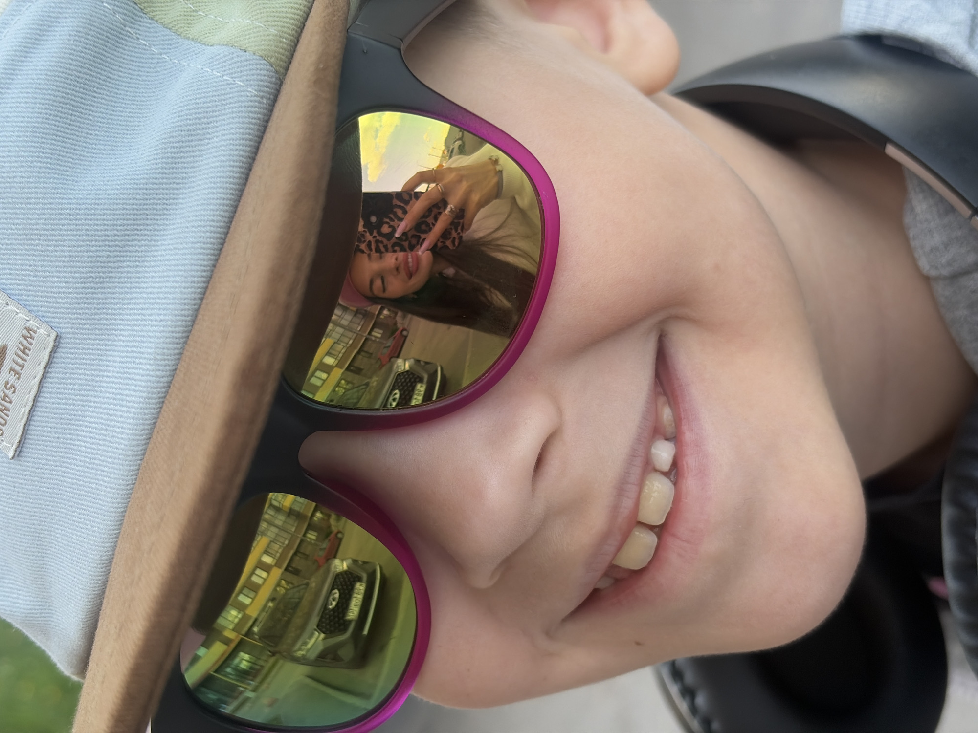 A close-up, rotated portrait features a young person, likely a child, smiling broadly. The subject's face is visible from just above the nose to the chin, revealing a wide, toothy grin. They are wearing sunglasses with distinctive purple frames and highly reflective, golden-green lenses. A light-colored bucket hat, possibly light blue and light green, is partially visible on the left side of the frame, with a tag bearing the text "WHITE SANDS." On the right side, a black headphone band can be seen, indicating the subject is also wearing headphones.
The reflective sunglasses offer two distinct scenes. In the lens closer to the viewer's right cheek, an older individual, likely a female, is reflected. She is holding a smartphone with a leopard print case, appearing to take a selfie. She has long dark hair, wears multiple rings on her fingers, and has long fingernails, showing a slight smile or smirk. In the other lens, a reflection of an urban environment is visible, featuring buildings and what appears to be a dark-colored SUV or truck, suggesting an outdoor city street or parking lot setting.
The bright reflections indicate a daytime, possibly sunny, setting. The overall mood conveyed by the subject's wide smile is one of happiness or playfulness. The location context for the image is Saint Petersburg, Russia.