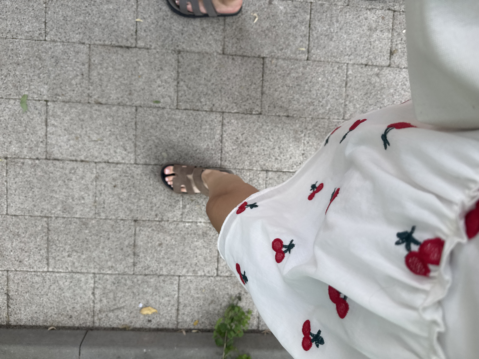 The image presents a top-down view of a paved outdoor area, capturing the feet of two individuals walking. In the immediate foreground, the lower leg and foot of one person are visible, clad in light brown, multi-strapped sandals. This person is wearing a white garment, likely a dress or skirt, which is embroidered with numerous red cherries, each featuring a dark green stem. The fabric of the dress appears ruffled and in motion.
Further back, the feet of a second person are partially visible, wearing dark grey or black sandals. Both individuals are traversing a sidewalk constructed from rectangular grey paving stones that exhibit a slightly textured, speckled surface. Scattered on the ground are small pieces of debris, including green leaves and a yellowish piece of litter. Along the very bottom edge of the frame, a dark grey curbstone separates the paved path from a narrow strip of earth and green foliage.
The lighting indicates it is daytime, though no harsh shadows are cast. The scene depicts a casual walk on a paved street or sidewalk, within the context of Plovdiv, Bulgaria. No text is visible within the image.