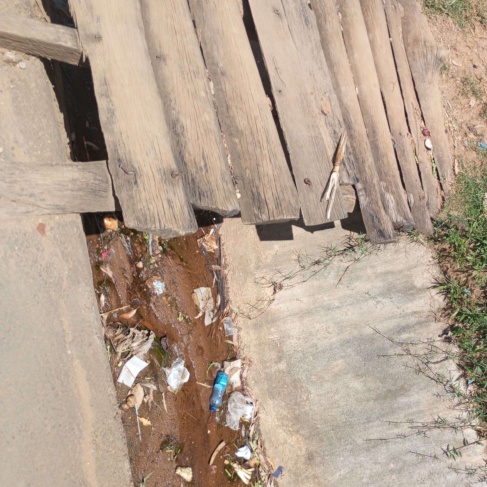 The image depicts a small wooden bridge over a concrete drain in Sheywe ward, Kenya. The bridge is constructed of weathered wooden planks, some with exposed nails. The drain is filled with stagnant water and littered with plastic bottles, plastic bags, paper, leaves, and other debris. The surrounding area is concrete with patches of grass and weeds growing along the edges. No people are visible in the image.