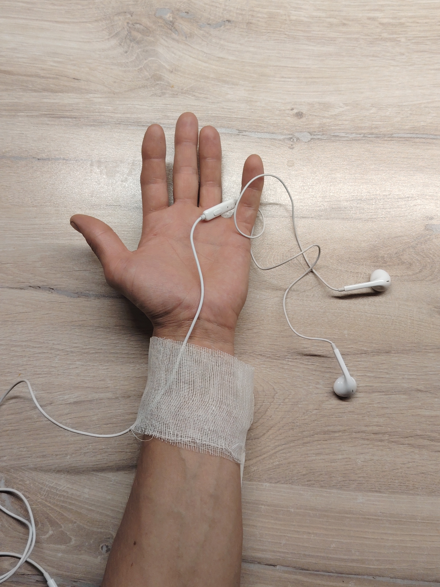 A high-angle shot shows a person's left hand and forearm resting on a light brown, wood-grain surface. A white bandage is wrapped around the wrist. White earbuds are draped across the hand and forearm. The location is unknown.