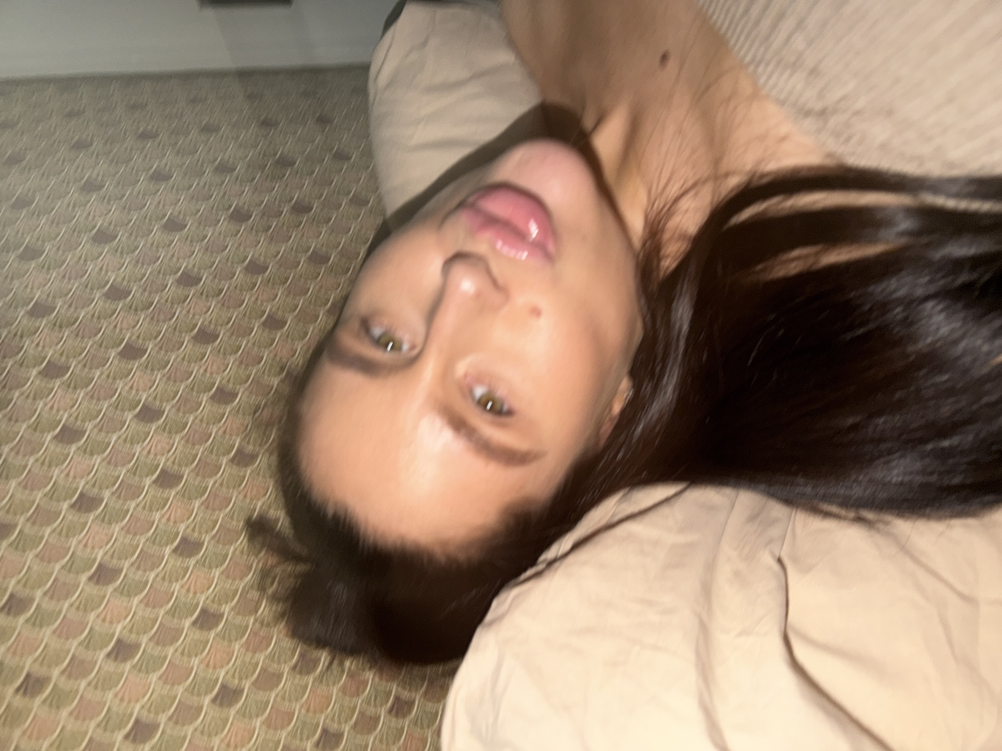 A woman with fair skin and dark hair is lying on a bed in a room with patterned carpet. The woman's head is tilted, and her lips are parted. The room appears to be indoors, and the carpet has a geometric pattern. No identifying information suggests the location is Khimki, Russia.