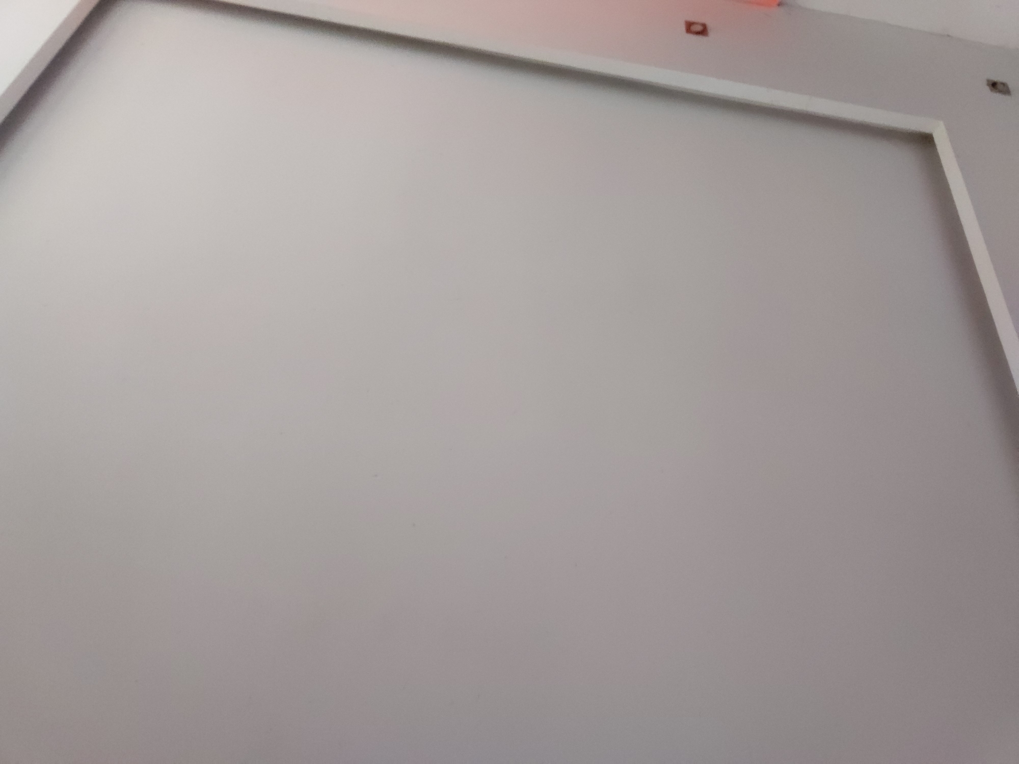 The image captures a light gray wall, presumably indoors in Santa Ana, Venezuela. A white decorative frame or molding outlines the top and right edges of the wall, creating a clean, geometric boundary. The wall surface is plain and unadorned. In the upper right portion of the image, above the white frame, a ceiling or upper section of the wall is visible, showing two small, dark, rectangular components that appear to be electrical outlets or fixture mounts. A faint reddish-orange hue or reflection is also present in this area, possibly from a light source. The lighting is even, indicating an interior setting. There are no individuals, activities, or visible text in the scene.