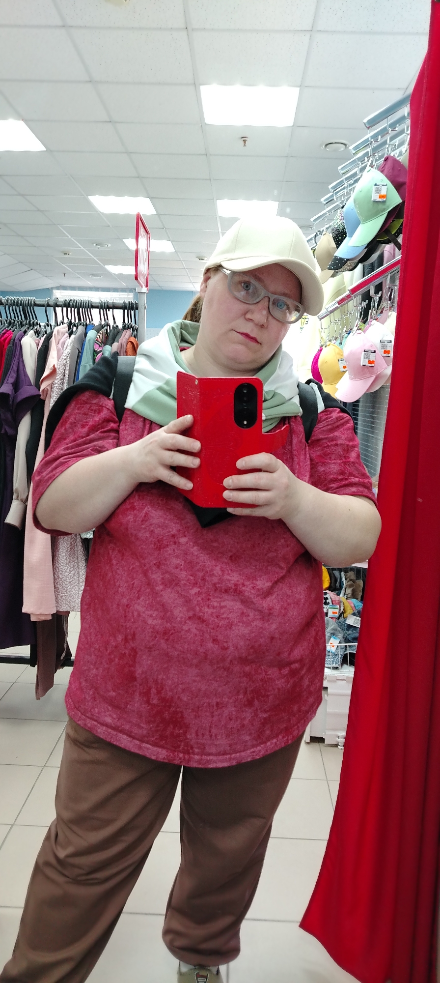 A woman stands in a shop, holding a red smartphone to take a photo in a mirror. She is wearing a light-colored cap, glasses, a red t-shirt, brown pants, and a green and white hooded scarf around her neck, with a black backpack visible. Racks of clothing hang in the background, along with a display of hats behind her right shoulder. A red curtain is visible on the right side of the image. The location is a store in Chaikovsky, Russia.