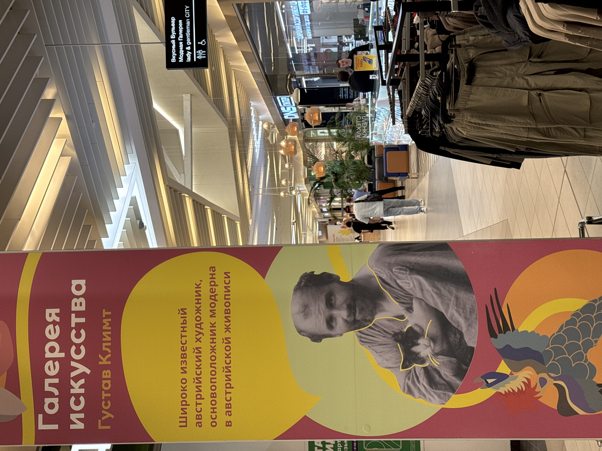 A medium shot of a shopping mall interior in Kazan, Russia, features a large promotional banner advertising a Gustav Klimt art gallery. The banner, positioned in the foreground, has images of the artist and his art. Behind it, a clothing rack displays neutral-colored pants. People are visible in the mall’s walkway, with shops and light fixtures extending into the background. Above, signage for "Lady & Gentleman CITY" and "Delicious Boulevard" are visible.