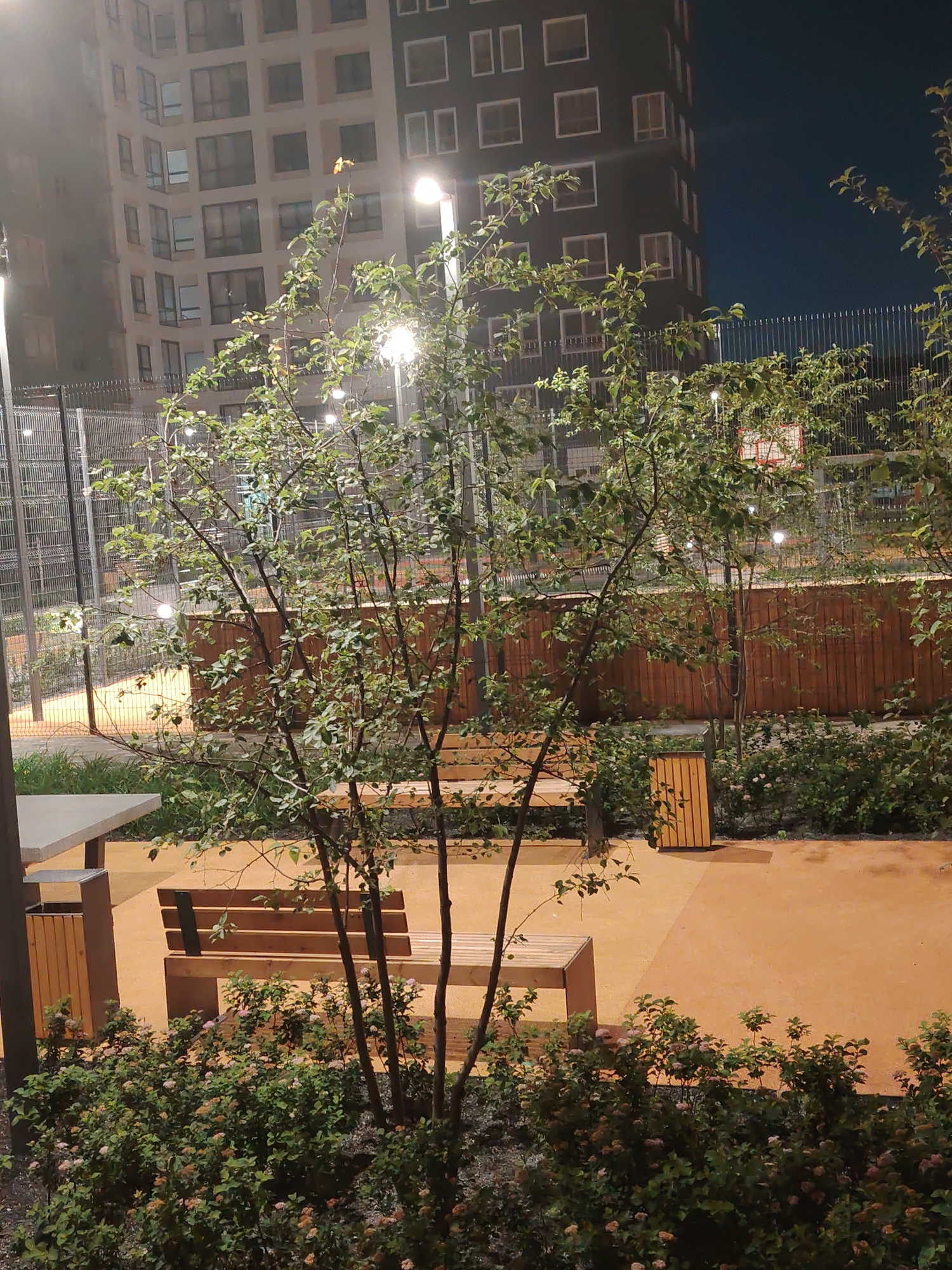 The image depicts an outdoor courtyard at night, likely in a residential complex in Yekaterinburg, Russia, given the style of the buildings. In the foreground, there are several young trees with green foliage, and a wooden bench with metal accents. To the left, a part of a concrete table and a trash bin with wooden slats are visible. The ground is covered in an orange-toned surface with some areas of grass and flowering bushes.
In the background, a tall, modern apartment building with numerous windows is illuminated. Behind this, a chain-link fence encloses a recreational area, possibly a sports court, with a basketball hoop visible. The scene is lit by artificial lights, including streetlights, and the sky is dark, indicating nighttime. The overall impression is of a well-maintained, urban public space designed for recreation and relaxation. There are no people visible in the image.