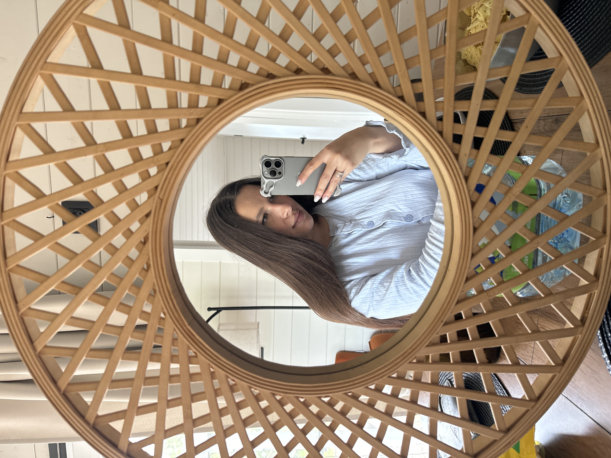 A woman is taking a selfie in a round, light brown, wooden-framed mirror. She is holding a smartphone with a silver case. The woman has long, brown hair and is wearing a light blue, button-down shirt. The background reflected in the mirror includes a white paneled wall, a floor, and various items, including what appear to be a lamp and some household objects. The location appears to be in an indoor setting, possibly a home. The city is Литвиново, Russia.