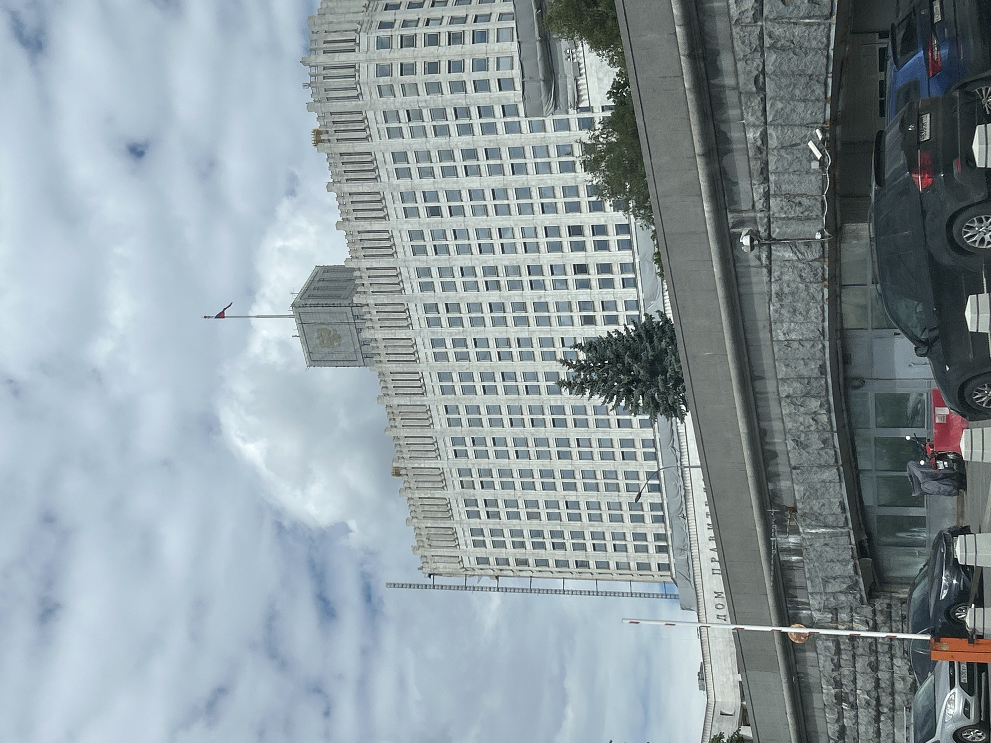 This image shows the White House building in Moscow, Russia, with the Russian flag flying from its rooftop. The building is a large, white, multi-story structure with many windows. In front of the building, there is a gray stone wall and a street with several cars parked. The sky is cloudy.