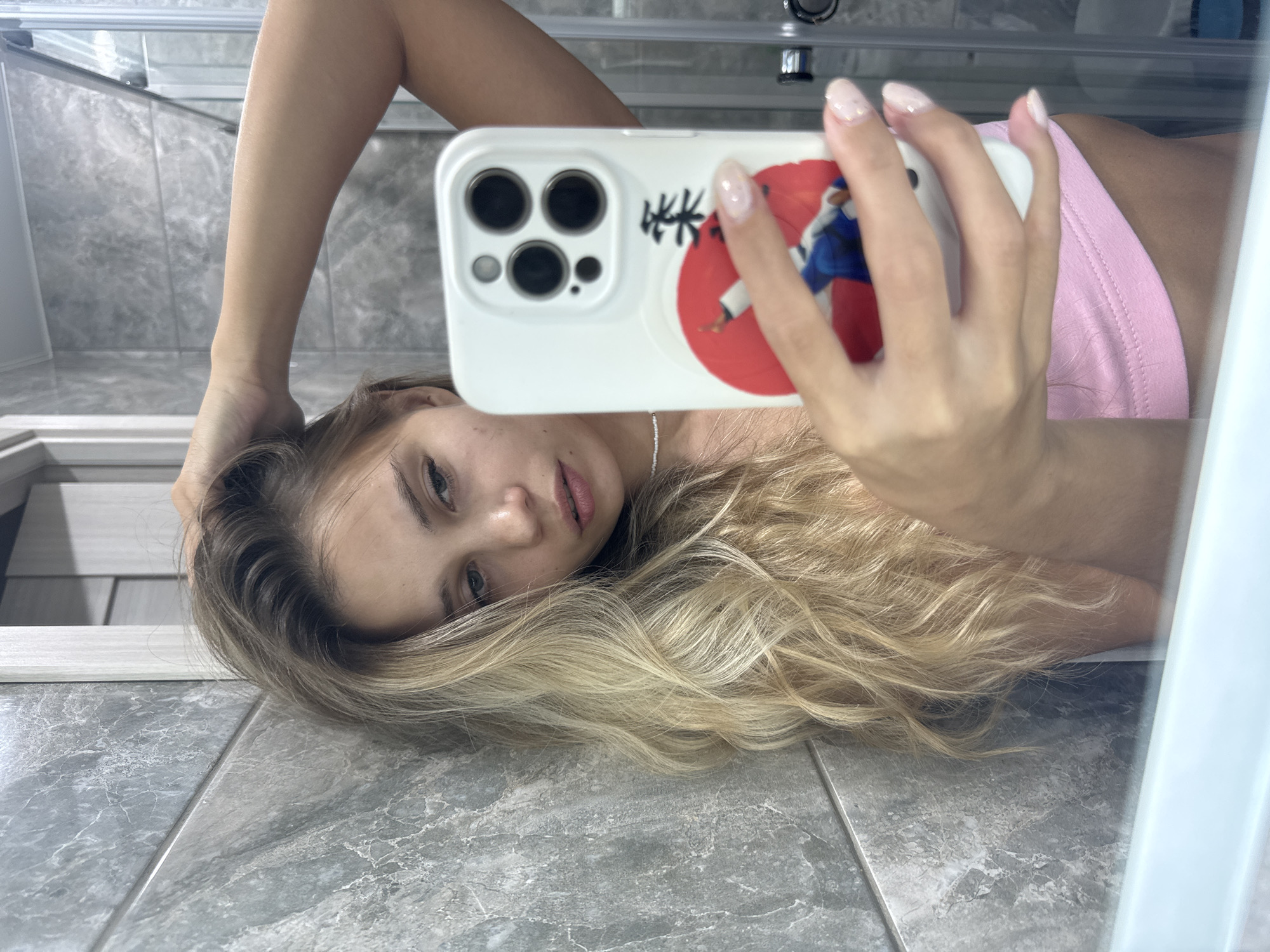 A young woman with long, blonde, wavy hair is taking a selfie in a bathroom. She is lying on a grey-tiled floor, with her head resting on the tiles and her left hand behind her head. Her right hand holds a white smartphone with a decorative case featuring a red circle and a figure in a blue and white uniform. She is wearing a pink top. The bathroom has a shower cubicle visible in the background. The location is likely in a residential building in Chaikovsky, Russia, given the interior design.