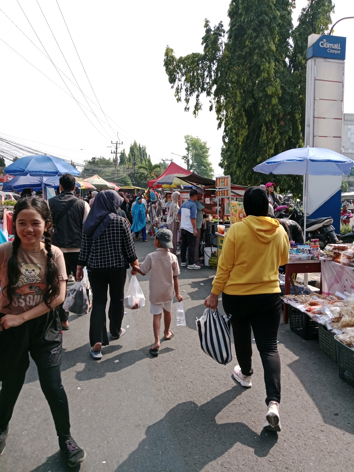 This is a photograph of a street scene in Cianjur, Indonesia, likely a market or a busy street with vendors. In the foreground, a young girl with braided hair smiles at the camera, wearing a brown t-shirt with a teddy bear graphic and black leggings. Behind her, people are walking in the street, some carrying shopping bags. To the right, a woman in a yellow hoodie and black pants walks away from the camera, carrying a striped bag. Further back, more people and market stalls with colorful umbrellas are visible. A sign for "Citimall Cianjur" is prominent on a tall pole in the background, indicating the location. The ground is asphalt, and there are shadows cast by the people and overhead structures. Electrical wires crisscross the sky.