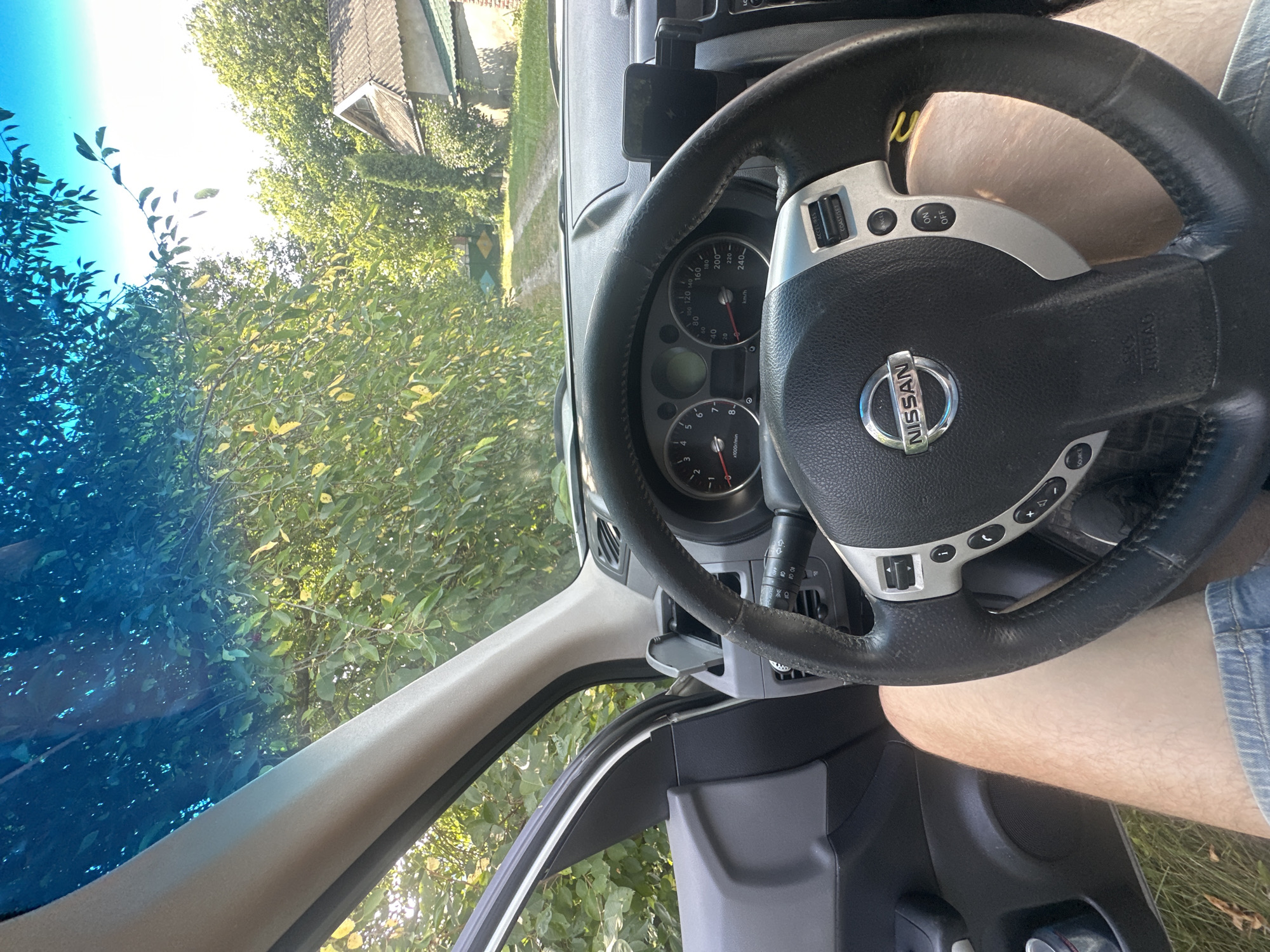 The image is taken from inside a Nissan vehicle, looking forward through the windshield. The steering wheel and dashboard are prominently visible, with the driver's legs in the foreground. Outside the car, a rural scene unfolds. Lush green trees line a dirt road that leads to a small, weathered house with a gray tiled roof. The sky is a clear blue, and the sun appears to be shining brightly. The overall impression is one of a quiet, rural setting. The image was captured in Shatava, Ukraine.