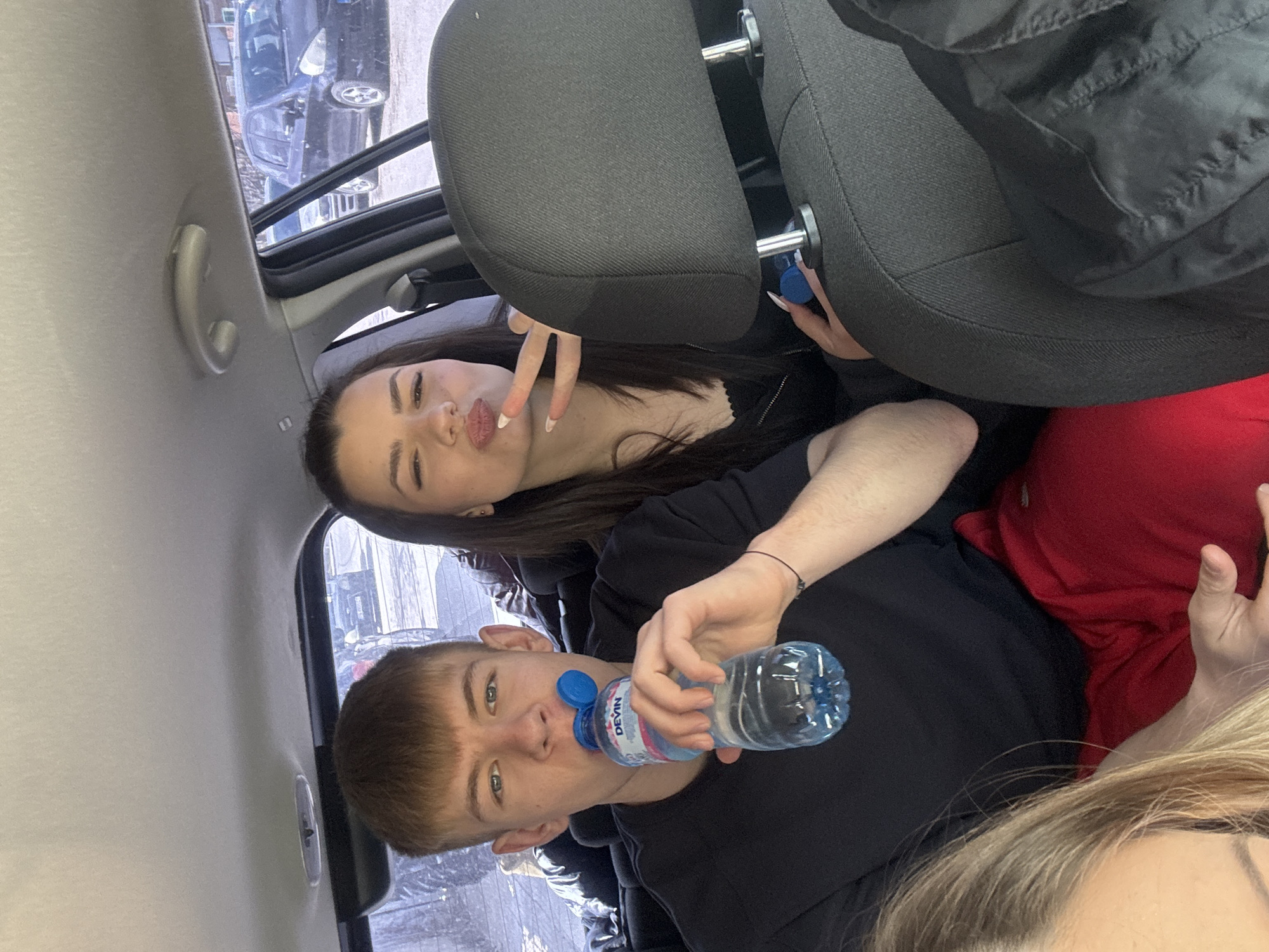 A group of young people are posing in a car interior. A young man is in the foreground, drinking from a plastic water bottle, while a young woman behind him is making a "peace" sign with her fingers. The environment outside the car window appears urban, potentially in Parvenets, Bulgaria. Other individuals are partially visible within the car, along with car seats and interior elements.