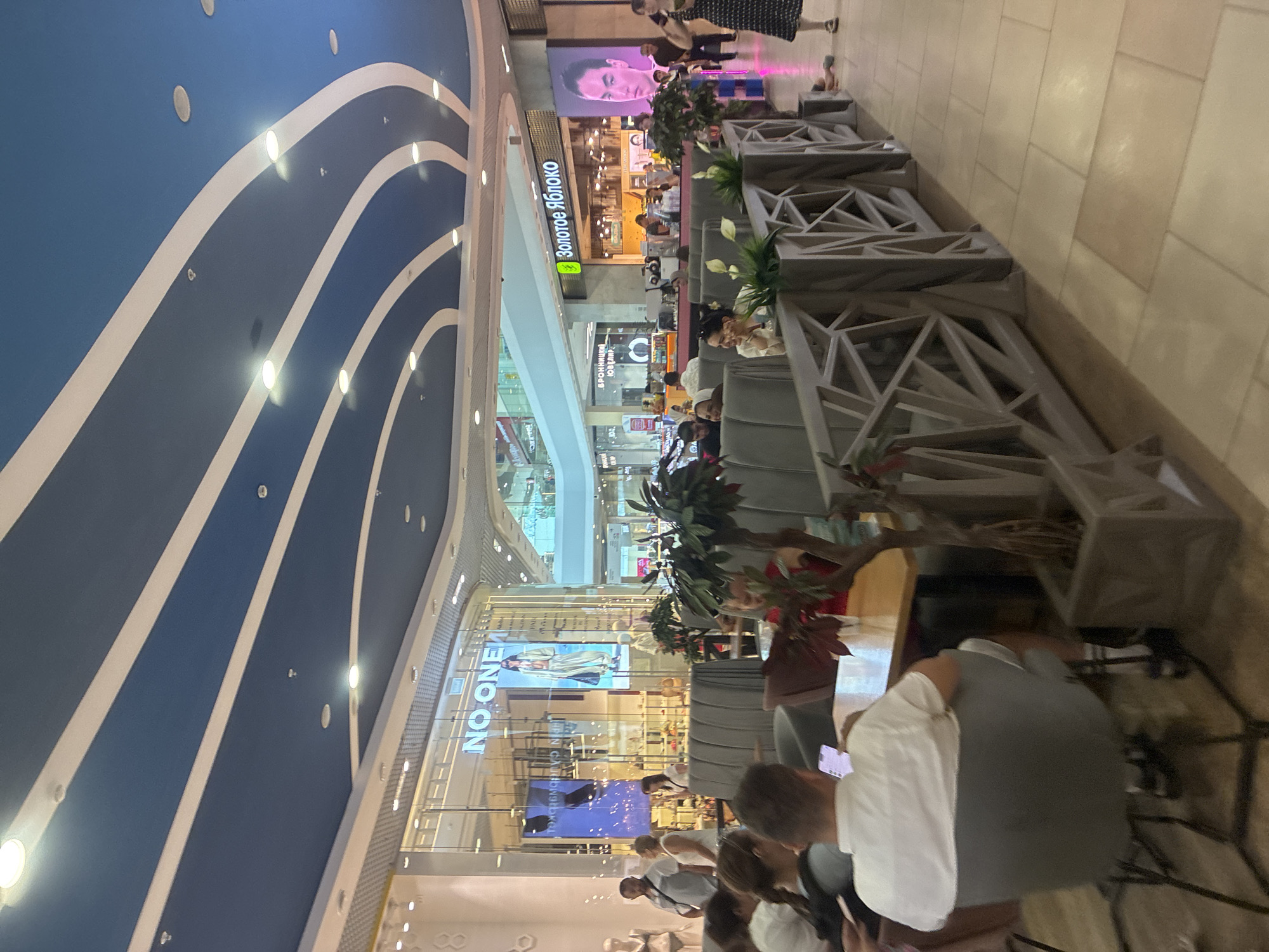 The image shows a spacious, modern interior of a shopping mall, likely in Sochi, Russia, featuring a distinctive blue and white striped ceiling with recessed lighting. In the foreground, a group of people are seated at tables, some engaged with their phones, in what appears to be a cafe or seating area with plush grey seating and decorative planters. To the right, a woman in a black and white polka dot dress walks by. Further back, there are storefronts visible, including one with the sign "NO ONE" and another with the Russian text "Золотое Яблоко." The overall atmosphere is one of a busy commercial space, with shoppers and patrons present.