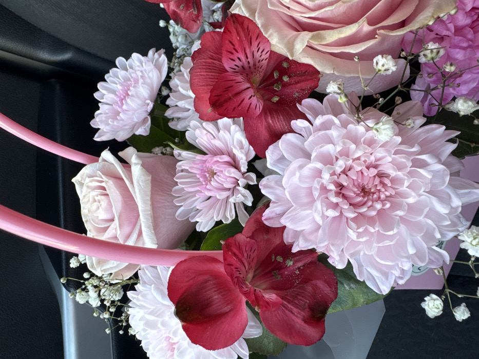 The image displays a close-up, top-down view of a floral arrangement situated within what appears to be a vehicle interior in Chaikovsky, Russia. The arrangement consists of a dense grouping of various flowers in predominantly pink, red, and white hues. Visible elements include several light pink roses, deep red alstroemeria with dark spotting on their petals, and numerous light pink and white chrysanthemums or similar composite flowers. Small white florets, consistent with gypsophila (baby's breath), are scattered throughout the arrangement. Two bright pink, curved, tubular structures, likely handles or support elements of a basket or box, are prominently positioned towards the center-top of the floral display. The background is dark, revealing a grey car door handle and a dark grey or black door panel on the upper left side, confirming an indoor, confined environment. No individuals, actions, or specific interactions are depicted.