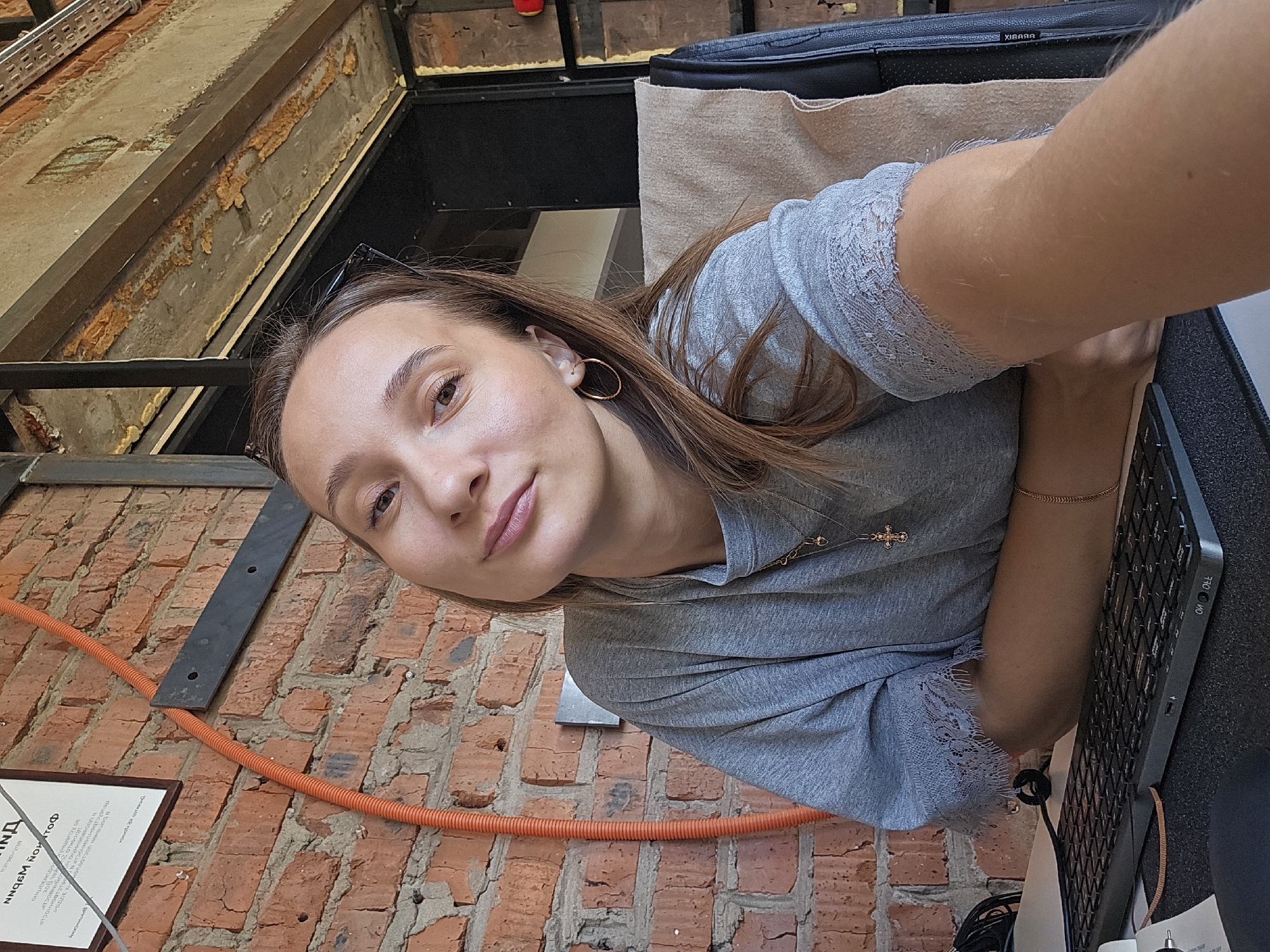 A young woman with light brown hair is sitting at a desk in front of a brick wall. She is wearing a gray t-shirt with lace trim on the sleeves and two gold necklaces. A pair of sunglasses is perched on top of her head. She is looking at the camera with a slight smile. In front of her is a black keyboard and part of a computer monitor. An orange conduit runs vertically along the brick wall behind her. The environment appears to be an office space with exposed industrial elements like metal beams and concrete. The presence of a diploma on the wall behind her suggests this may be an office of educational or professional certification in Chaikovsky, Russia.