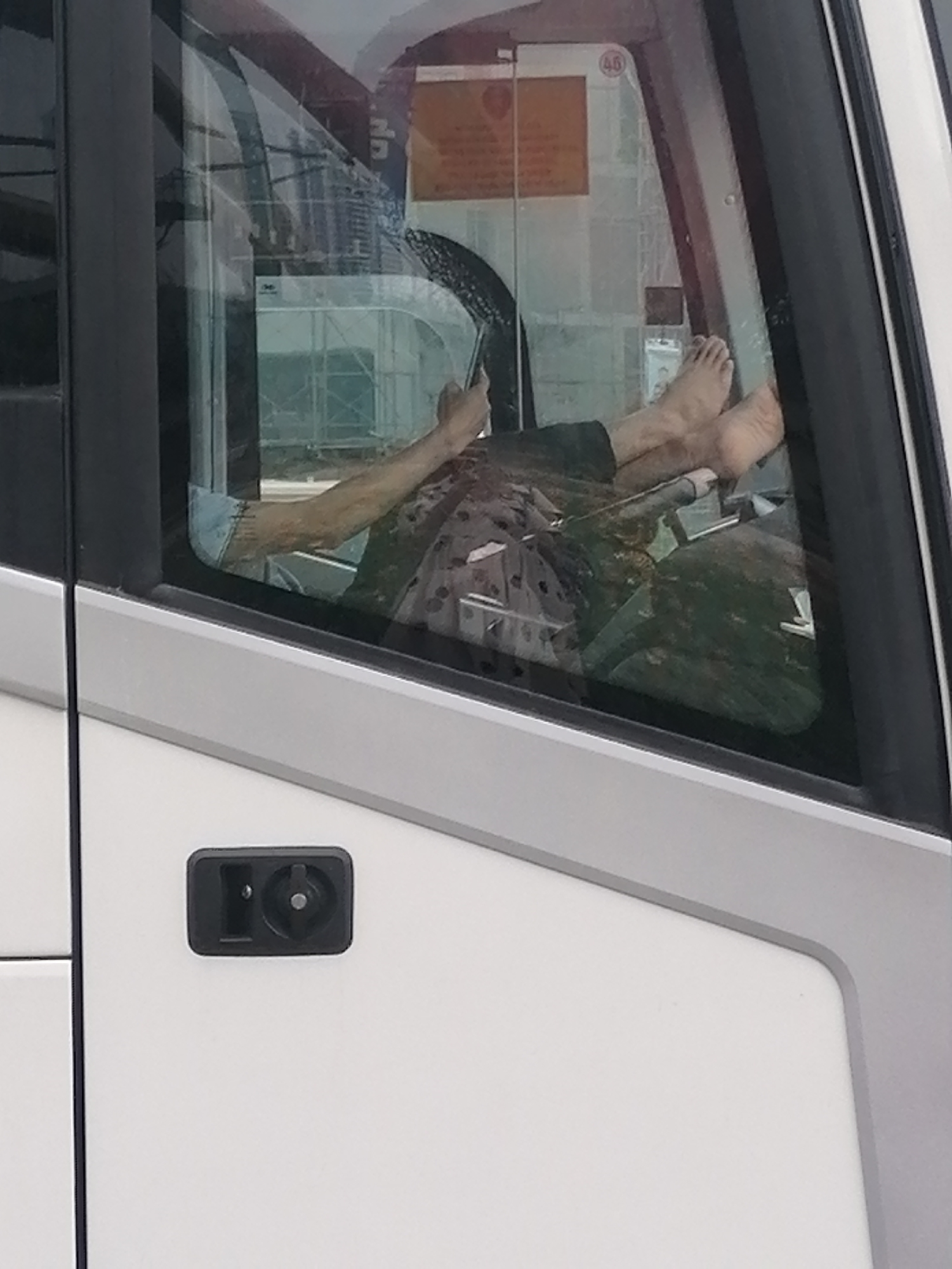 The image captures a partial view of the interior of a vehicle, likely a bus or coach, seen from the outside through a side window. A person is visible inside, relaxing with their bare feet propped up on what appears to be the dashboard or control panel. One of the person's arms is also visible, resting.
Through the transparent window, an outdoor scene with metal scaffolding is discernible, suggesting a construction site or nearby building. The vehicle's window is framed by dark grey and light grey elements, with a white lower door panel featuring a black handle or latch.
A rectangular orange sign is visible through the window, positioned inside the vehicle. The sign has a circled number "46" in its top left corner, but other text on the sign is illegible due to blur and distance. The scene is illuminated by ambient daylight, indicating either an overcast day or an indoor setting with natural light. The relaxed posture of the person suggests they are resting or waiting. The context for this scene is Bai Chay Ward, Vietnam.
