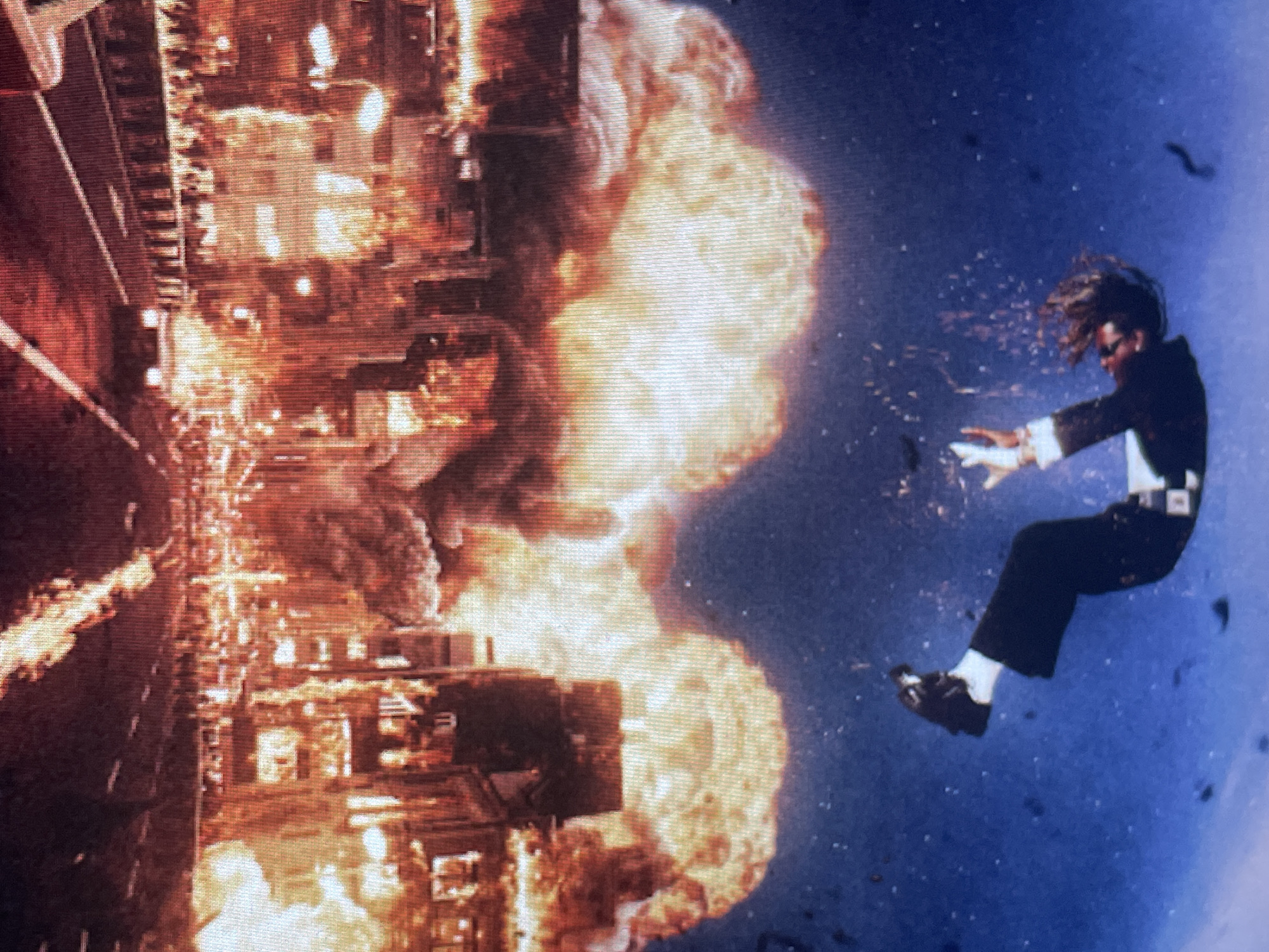 This media file captures a dramatic, action-packed scene, likely from a film or music video, depicted with the image rotated 90 degrees counter-clockwise. When viewed upright, the scene unfolds with a massive explosion engulfing an urban landscape at the bottom and a person being thrown through the air against a dark sky at the top.
At the bottom of the frame, a cityscape is consumed by an immense conflagration. Bright orange and yellow flames, accompanied by plumes of dark smoke, erupt from multiple structures, suggesting widespread destruction and collapsing buildings. Fragments and debris are visible within the fiery inferno. What appears to be a road or an elevated structure runs along the bottom-left, showing signs of damage from the blast.
In the upper portion of the scene, against a deep blue, almost purplish sky, a male figure is suspended in mid-air. He is dressed in a formal attire: a dark suit jacket, a white shirt with ruffled cuffs, dark trousers, and dark shoes paired with white socks. His dark hair is dramatically swept back, indicative of intense motion or wind. He wears sunglasses, obscuring his eyes, and his arms are outstretched, hands open, suggesting he is caught in the force of an explosion or fall. Small particles or debris are scattered around him in the air.
The activity depicted is a catastrophic event, likely a large-scale explosion, with the individual being violently propelled away from the blast. The stark contrast between the bright, fiery explosions and the dark blue sky suggests the event takes place at night or during twilight. No discernible text is visible within the image itself. The location context provided is Sofia, Bulgaria. The overall impression conveyed is one of chaos, extreme danger, and high-stakes action.