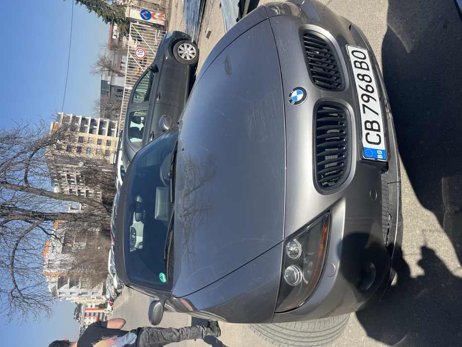A dark grey BMW Z4 convertible (E85 generation) with a black soft top is parked outdoors on a paved surface under clear blue skies in Sofia, Bulgaria, identifiable by the "CB" designation on its front license plate, "CB 7968 BO". The car's hood exhibits surface dust or marks and reflects bare tree branches. A green sticker is affixed to the upper left corner of the windshield.
To the left of the BMW, a person is partially visible from the mid-torso down, wearing dark jeans and a dark t-shirt with a colorful graphic on the back. They stand facing away from the camera, casting a shadow on the ground alongside the car's shadow.
In the mid-ground, a dark grey sedan is parked behind and to the right of the BMW. Further right, a white metal fence or gate features a red circular sign displaying the number "10" and a smaller blue directional sign.
The background consists of several multi-story buildings with light-colored facades and numerous windows, indicative of an urban or residential area. Bare-branched trees are scattered throughout the scene, and a line of additional parked vehicles is visible in the distance. The scene is brightly lit by natural sunlight, casting distinct shadows.