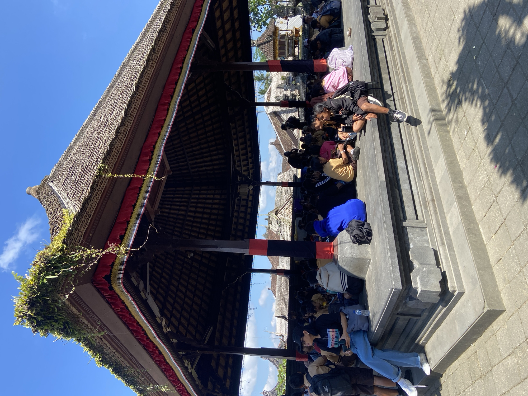 This image depicts an outdoor scene at the Kehen Temple in Bangli, Bali, Indonesia, under a clear blue sky with scattered clouds. A large, open-air pavilion with a multi-tiered, thatched roof dominates the foreground and midground. The roof's edge is adorned with decorative red, yellow, and black fabric trim, and vines with green foliage cascade from the top. Inside the pavilion, a group of people, appearing to be mostly young students in uniforms or casual attire, are seated on the raised stone platform. Some are interacting with each other, while others are looking around or resting.
In the background, the distinctive architecture of the Kehen Temple complex is visible, characterized by stone structures and thatched roofs, characteristic of Balinese temples. The ground surrounding the pavilion is paved with stone tiles, and shadows from trees or structures are cast across it. The overall atmosphere suggests a gathering or a break during an excursion or educational visit.