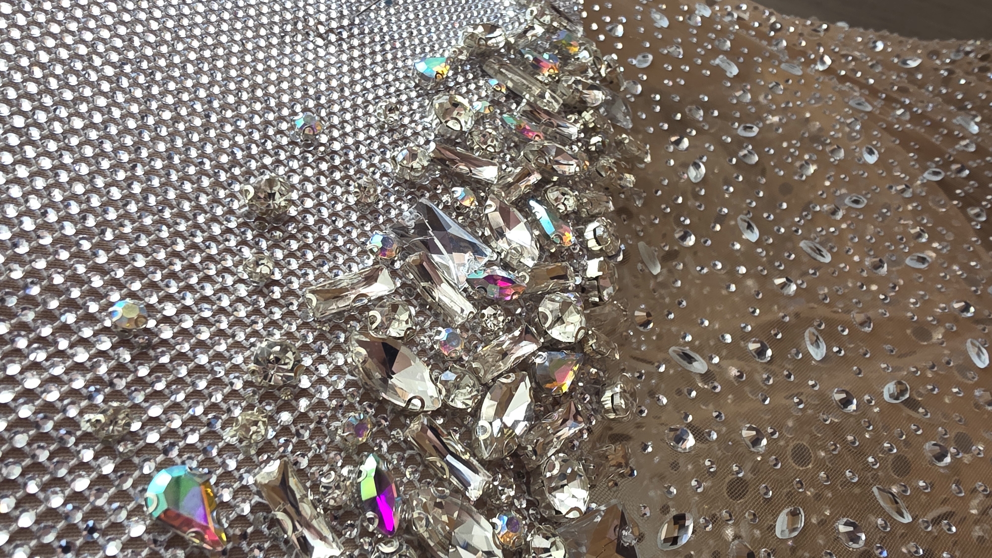 The image shows a close-up of a garment embellished with various sizes and shapes of clear crystals and rhinestones. The background fabric varies; on the left, it is a dense, tightly woven mesh covered in small, uniformly sized rhinestones. In the center, larger, irregularly shaped and sized crystals are densely clustered. On the right, the background fabric is a more open mesh with a sparse scattering of small, oval-shaped rhinestones. Iridescent reflections are visible on several of the crystals. The location is not identifiable from this image.