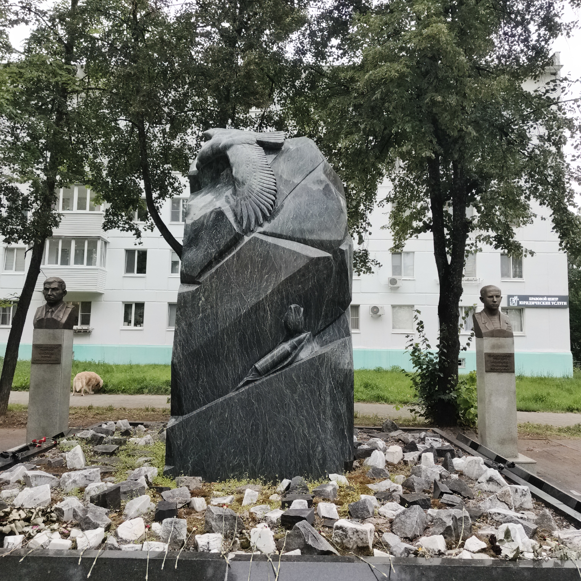 This media file depicts a memorial monument in Chaikovsky, Russia. The monument itself is a large, dark stone sculpture featuring an eagle in flight and a projectile, possibly a bomb or missile, carved into its surface. It is surrounded by a scattering of broken stones and some scattered flowers, suggesting a somber or commemorative purpose.
To the left and right of the central monument, there are two busts of men on stone pedestals. The bust on the left appears to be of a man in military attire. A plaque is visible on its pedestal, but the text is too small to read clearly. To the right, another bust of a man is visible, also with a plaque, and above this bust, a sign reads "ПРАВОВОЙ ЦЕНТР ЮРИДИЧЕСКИЕ УСЛУГИ" (Legal Center Legal Services).
In the background, a multi-story apartment building with white walls and numerous windows is visible. Trees with green foliage flank the monument and the building. A light-colored dog is seen in the middle ground, sniffing the ground near the base of the left pedestal.
The scene appears to be outdoors, likely in a public space or park area adjacent to residential buildings. The lighting suggests it is daytime, possibly overcast, as there are no strong shadows. The overall atmosphere is solemn, given the nature of the monument and the scattered stones.