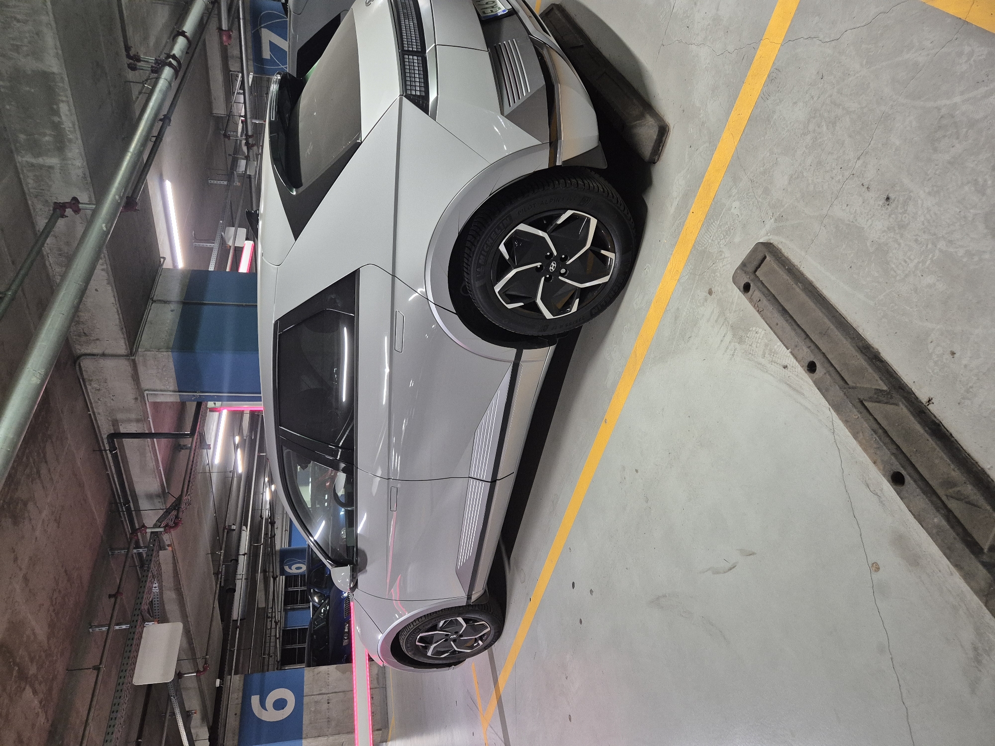 A silver Hyundai Ioniq 5 electric vehicle is parked in an indoor parking garage. The car is positioned at an angle, facing towards the right of the frame, with its rear visible. The parking garage has a concrete ceiling with exposed pipes and conduits. Fluorescent lighting illuminates the space. Blue and white markings with the number "6" are visible on a pillar in the background, indicating parking bay number 6. Yellow lines are painted on the concrete floor, marking parking spots. A dark-colored, textured bumper stop is placed behind the car's rear wheels. The overall environment appears to be a typical urban parking facility. The license plate of the vehicle is partially visible and appears to be Bulgarian, with characters "EA61". The image was taken in the parking facility of the Paradise Center mall in Sofia, Bulgaria.
