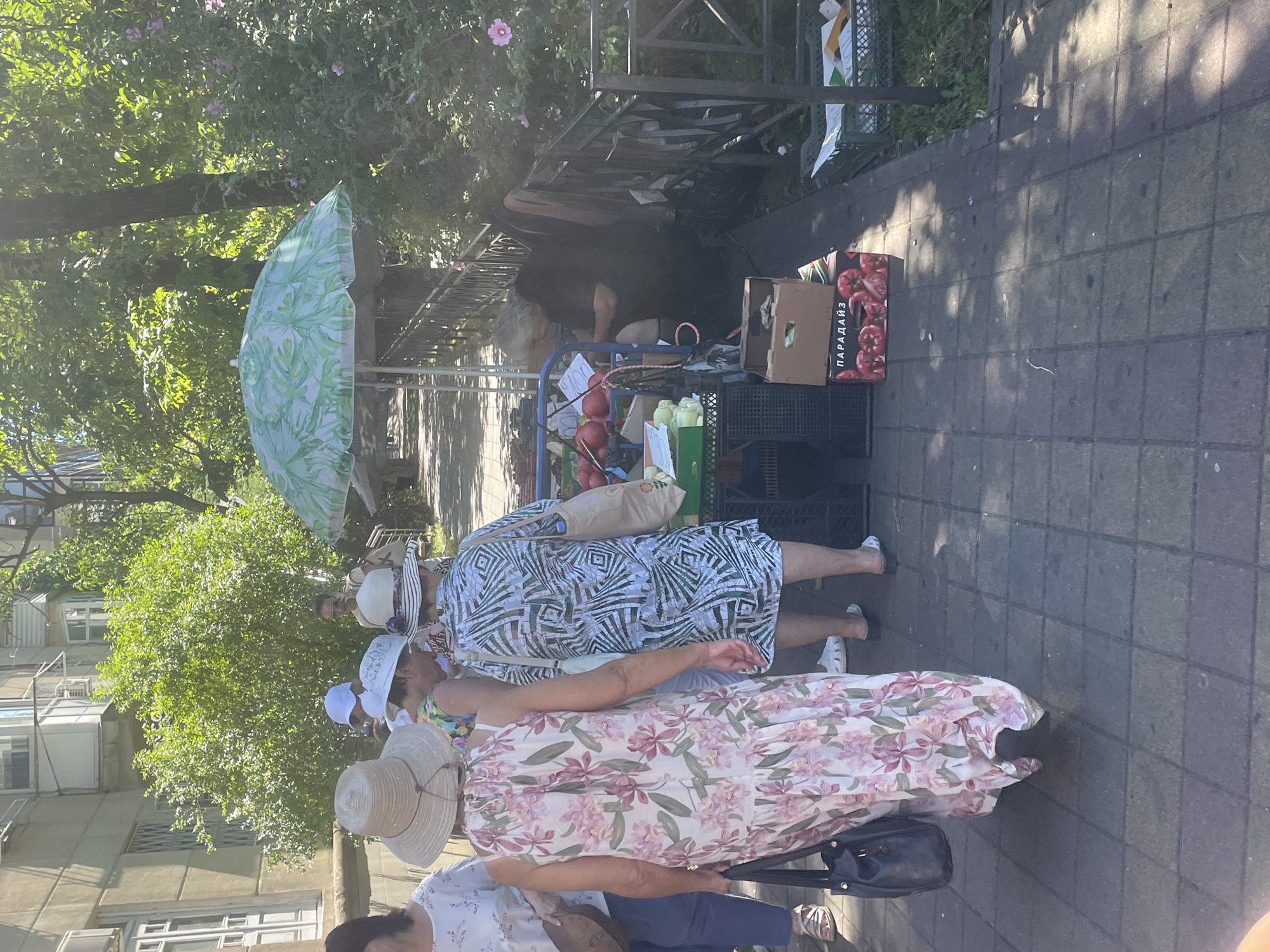 The image depicts an outdoor market scene on a paved walkway, likely in Sochi, Russia, given the context of the request. In the foreground, three women are visible from the waist down, two of them wearing wide-brimmed hats. One woman on the left wears a long, floral-print dress, and the woman next to her is dressed in a patterned geometric-print dress. Both are facing away from the camera. A third woman in a white hat is partially visible behind them.
Behind the women, a market stall is set up under a green and white leaf-patterned umbrella. The stall displays an assortment of produce, including red tomatoes in a cardboard box labeled "ПАРАДАЙЗ" (Paradise) in Russian. To the right of the stall, a person in a dark outfit appears to be seated and possibly engaged with a phone, partially obscured by the stall's structure and the surrounding greenery. Green crates and other market items are visible throughout the scene. The environment suggests a sunny day, with dappled sunlight filtering through the trees that line the walkway. The architecture in the background appears to be residential buildings.