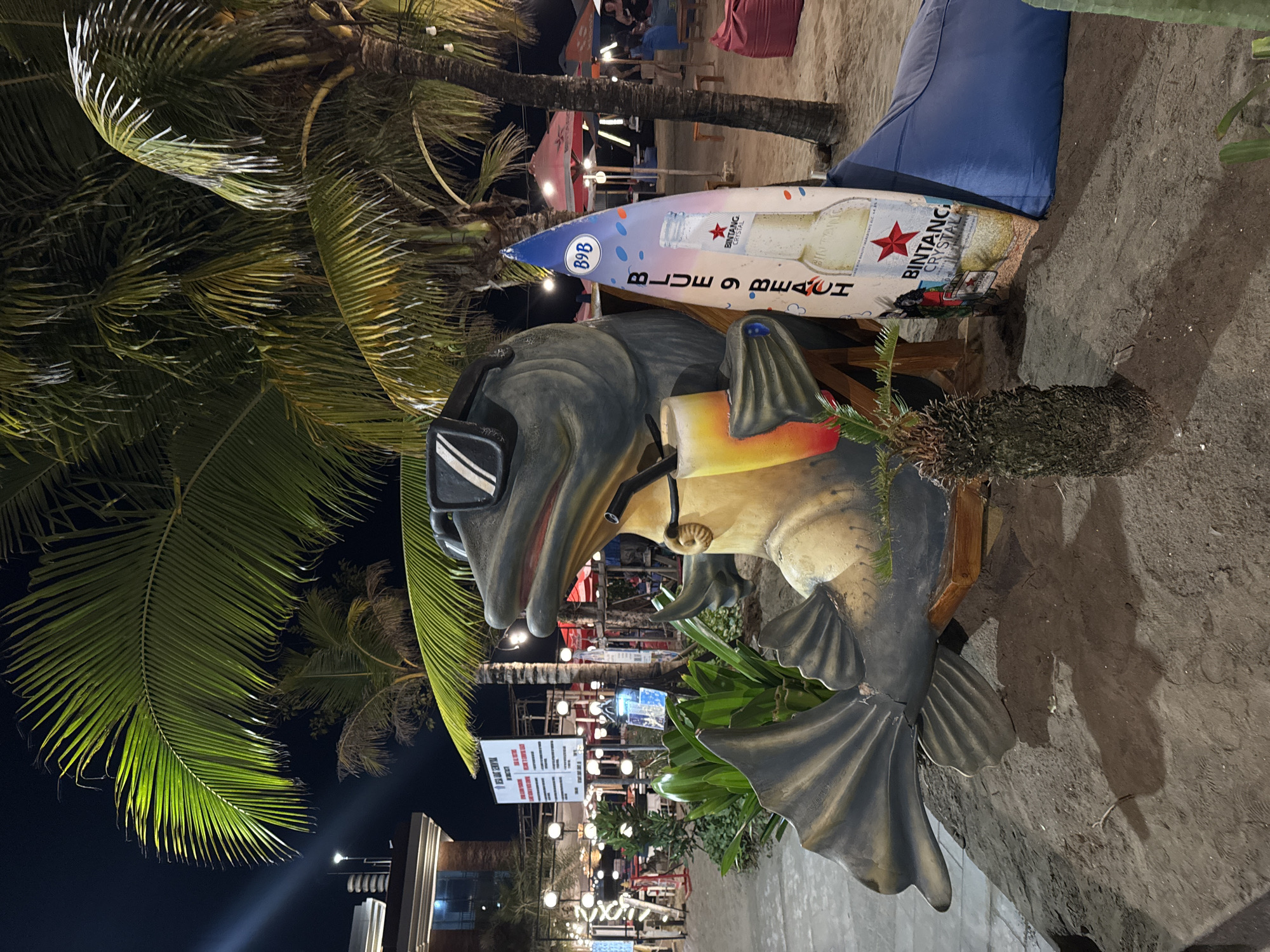 At night on a sandy beach in Kuta, Indonesia, a large, stylized fish sculpture wearing sunglasses sits in a wooden chair. A drink with a straw rests in its hand, and a surfboard leaning against it displays a "Blue 9 Beach" advertisement and a picture of a Bintang beer bottle. Palm trees and string lights illuminate the scene, with beach umbrellas and seating areas visible in the background.
